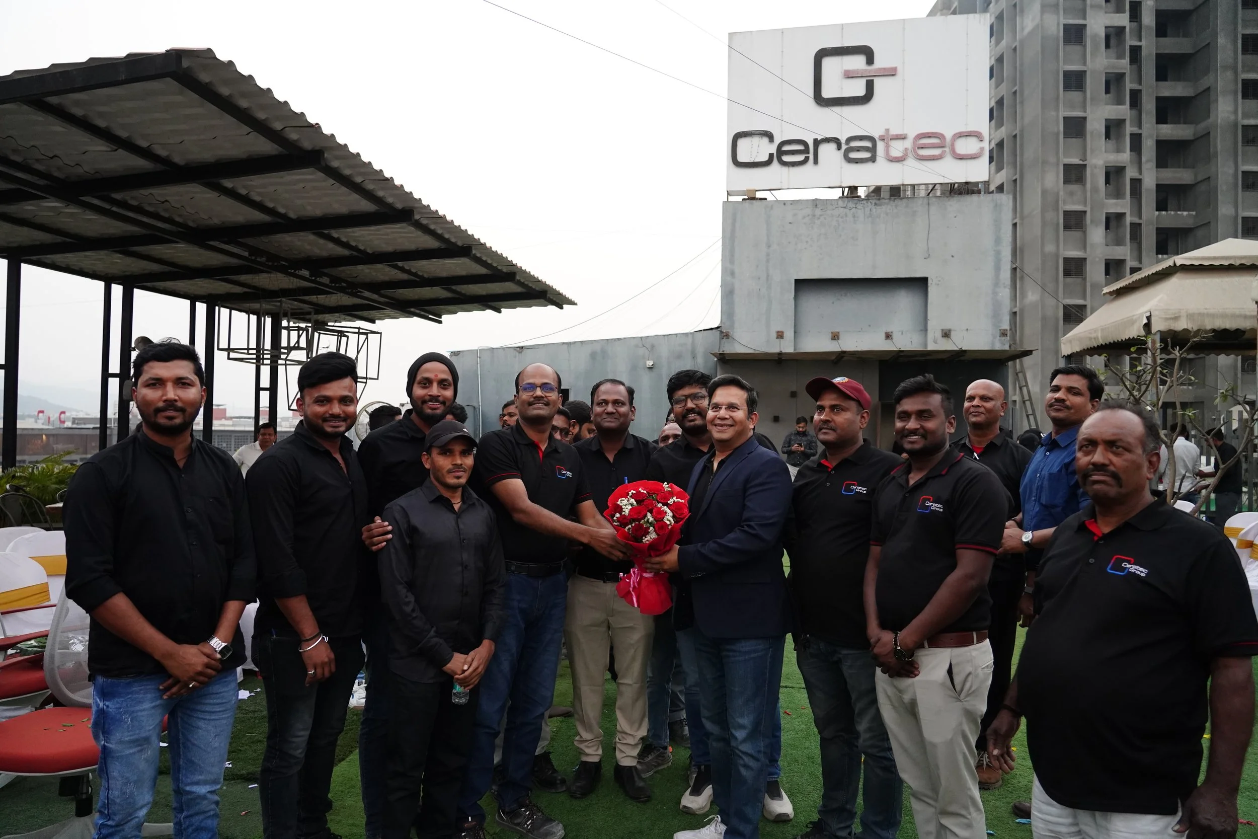 Group of men in black shirts gathering on a rooftop, with one man presenting a bouquet of flowers to another man in a navy blazer. The rooftop has seating and tiki torches, and a sign reading 'Ceratec' is in the background.