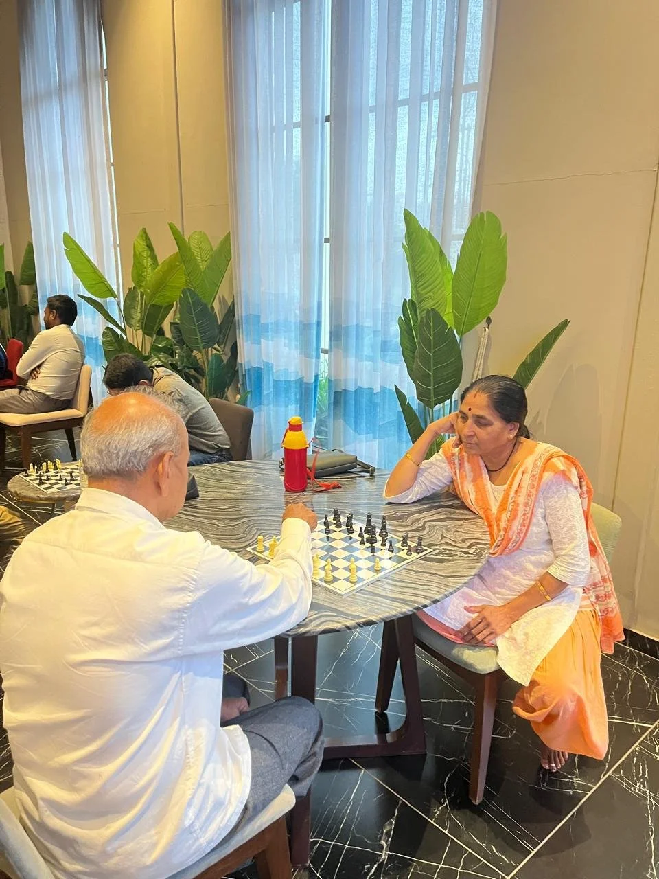 Two people playing chess at a round table in a room with large potted plants and sheer curtains covering tall windows.