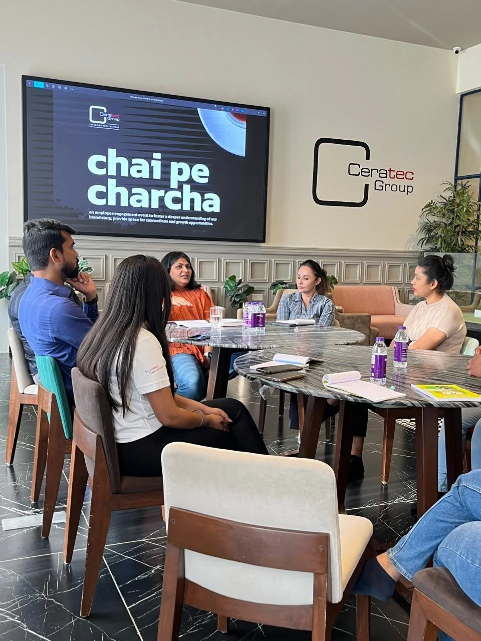 Group of people sitting around a table having a discussion in a modern conference room with Ceratec Group branding and a large screen displaying a presentation.