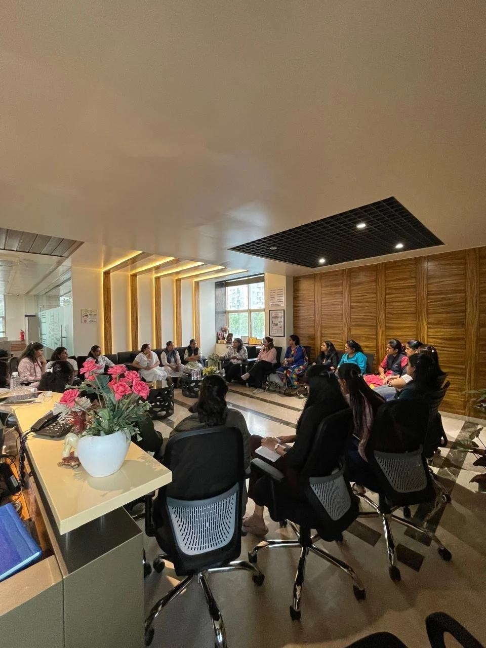 A group of women seated in a circle in a modern conference room, engaging in a discussion. The room has wooden paneling, large windows, and a table with pink flowers in a white vase.
