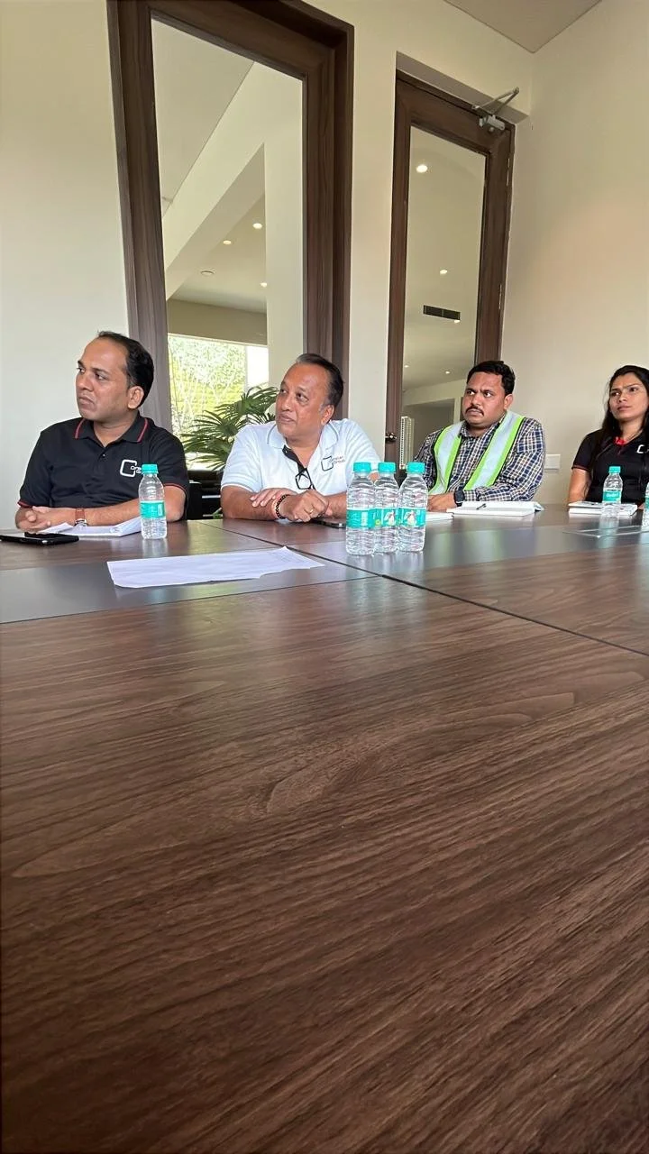 Four people sitting at a wooden conference table with water bottles in front of them inside a room with large windows.
