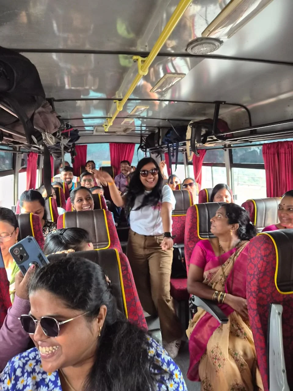 People on a bus smiling, some wearing sunglasses, with a woman standing and dancing in the aisle. Red curtains, luggage above, and a window showing part of the outside scenery.