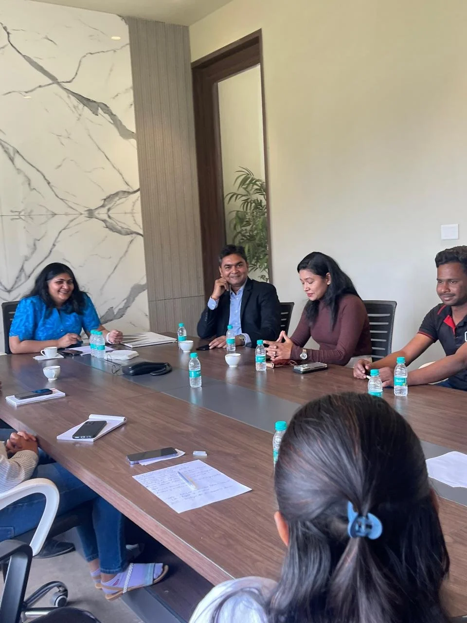 Meeting with five South Asian professionals seated around a large conference table, with notebooks, smartphones, pens, water bottles, and cups on the table, in a modern office with marble and wood wall decor.