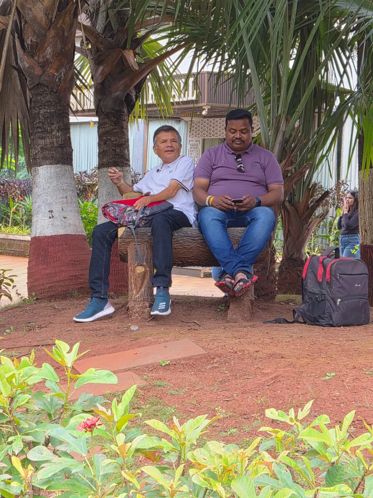 Two men sitting on a wooden bench under large palm trees. One man is talking, and the other is looking at his phone. There are backpacks nearby, and a woman is in the background.