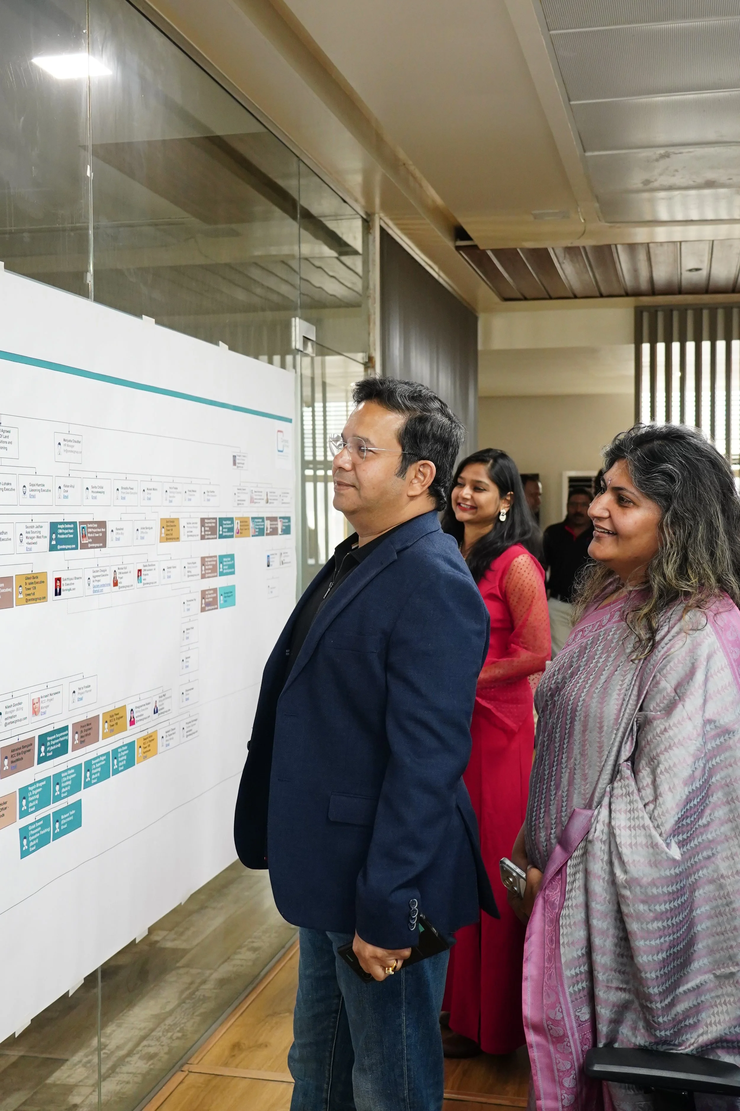 Three people observing a detailed chart or diagram on a wall.