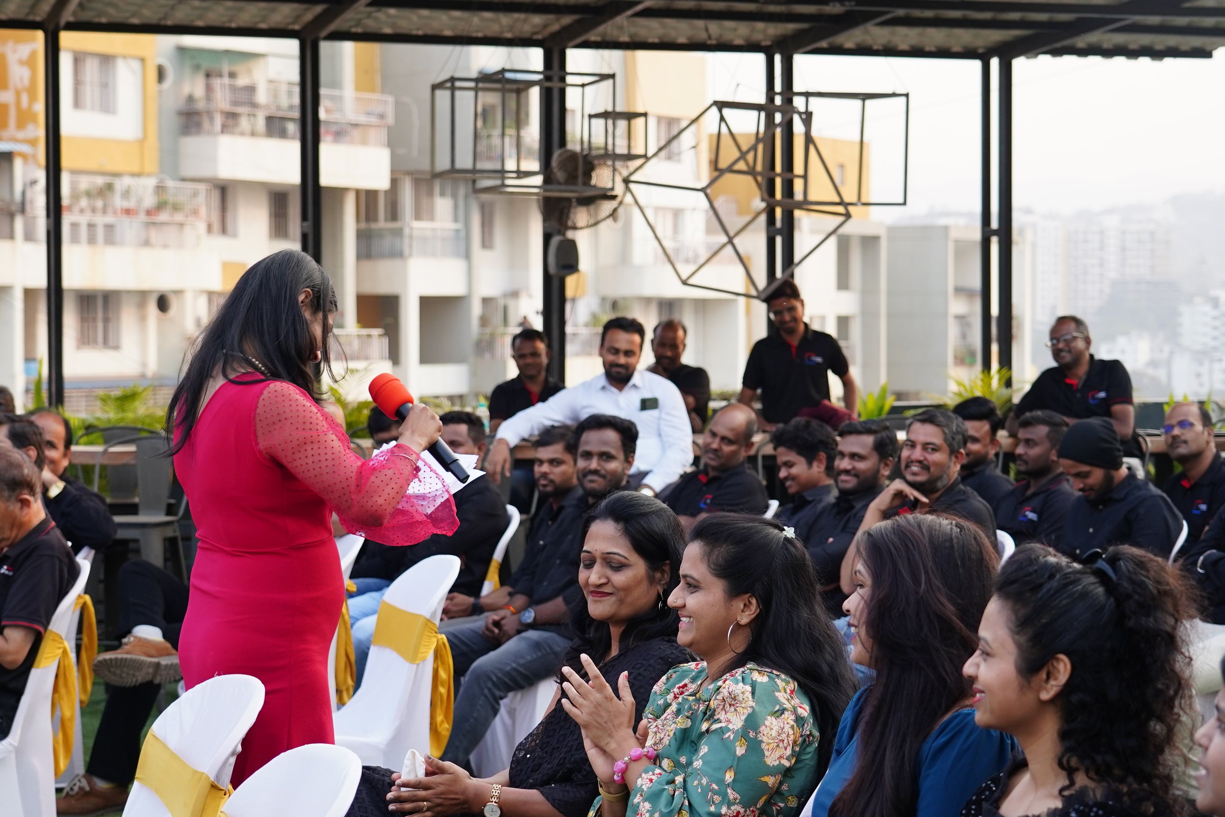 A woman in a red dress with sheer sleeves standing and speaking into a red microphone to an audience sitting on white chairs with gold sashes, on a rooftop with city buildings in the background.