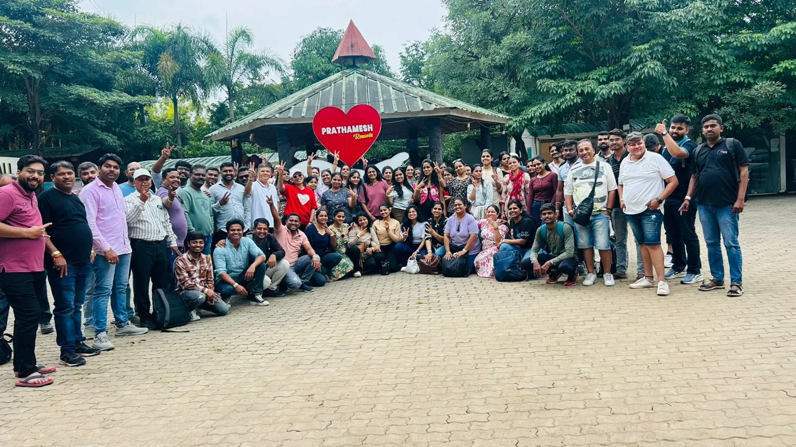 A large group of people gathered outdoors, posing for a photo in front of a red heart-shaped sign that says "PRATHAMESH Recurve". The group includes men and women of various ages, some making peace signs, and they are standing on a paved area surrounded by green trees.