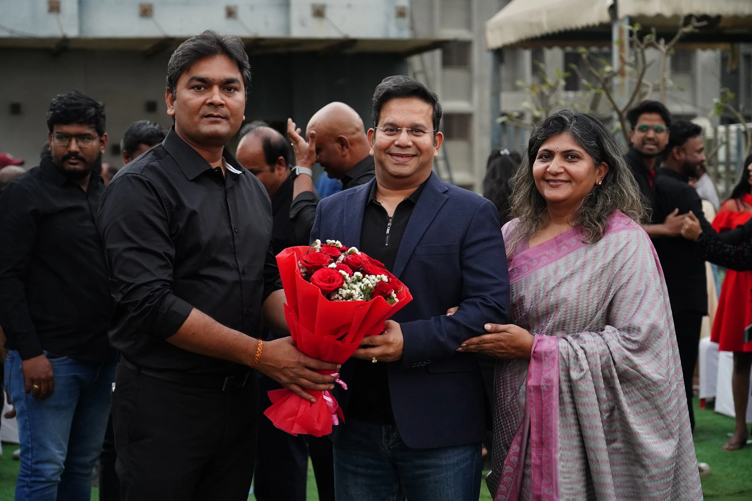 A group of three people, two men and one woman, standing together at an outdoor event, with the man in the middle holding a bouquet of red roses wrapped in red paper, smiling at the camera, surrounded by other people in the background.