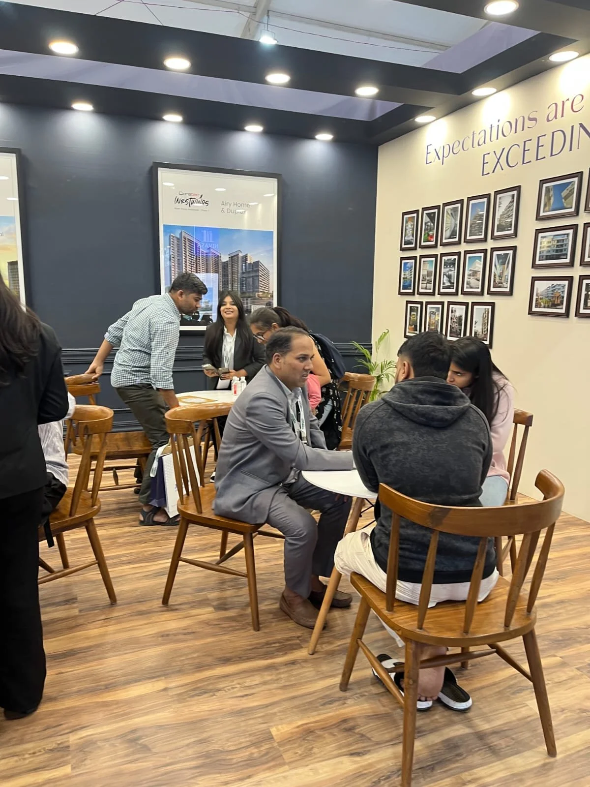 People sitting and talking at small round tables in a modern indoor space with wood flooring and framed photos on the wall. Some people are standing and engaging with each other, while others are seated in casual and business attire.