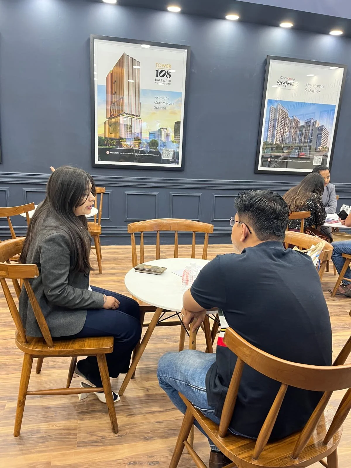 Two people seated at a round table engaged in conversation inside a cafe or restaurant. The woman has long dark hair and is wearing a blazer, while the man has short dark hair, wearing glasses and a black T-shirt. The background features a dark blue wall with framed advertisements for real estate developments and several other couples seated at tables.