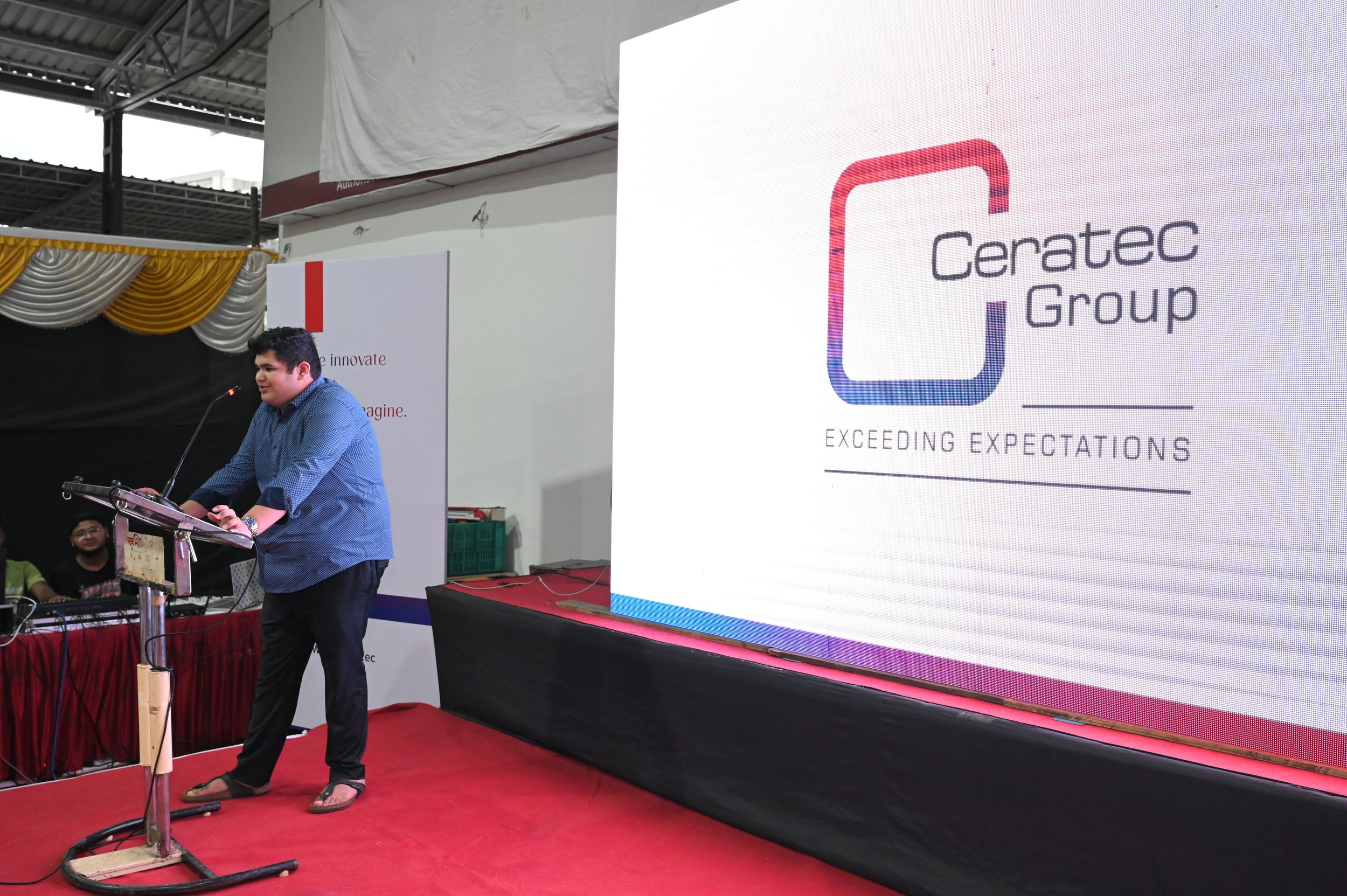 A man in a blue shirt and sandals standing at a podium giving a speech during a corporate event. Behind him is a large digital screen displaying the Ceratec Group logo, tagline 'Exceeding Expectations,' and a white backdrop with a red stripe.