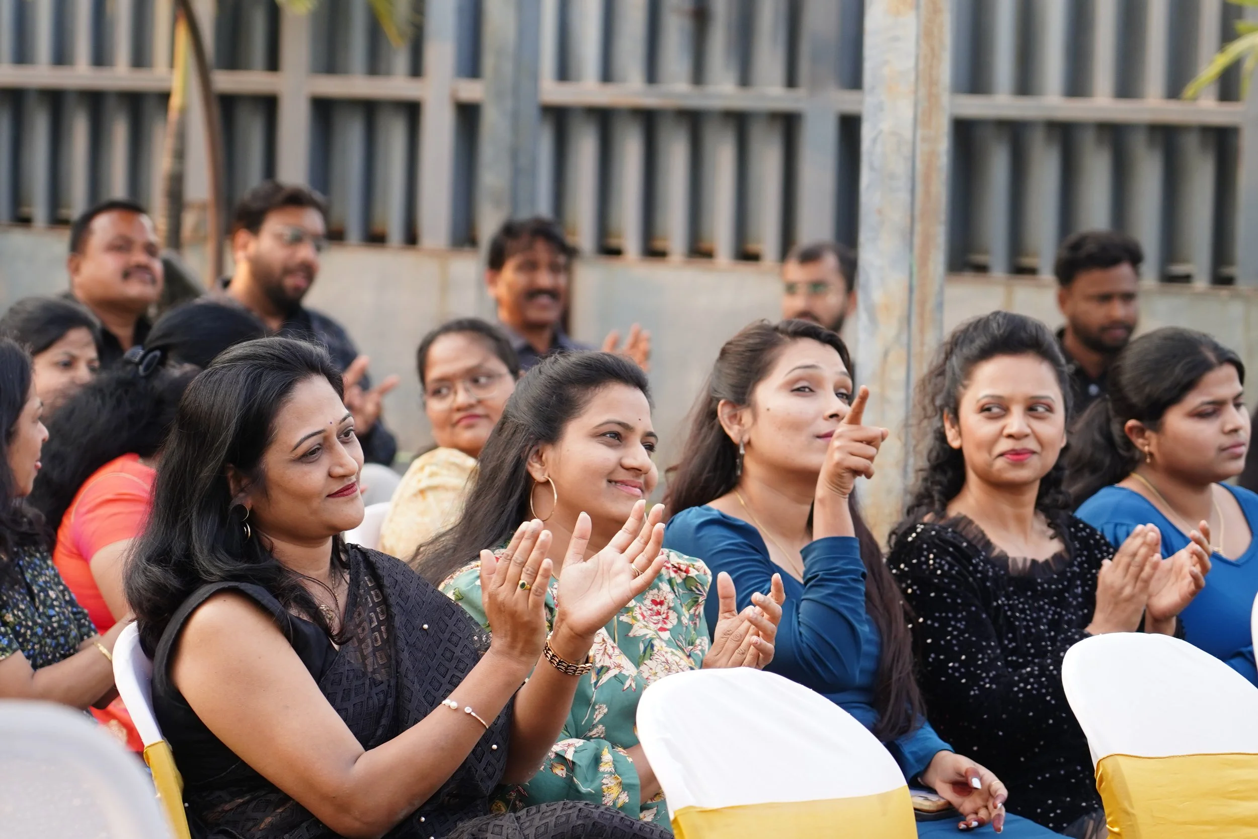 A group of women and men sitting outdoors, attending an event, some clapping and smiling, in front of a metal fence and plants.