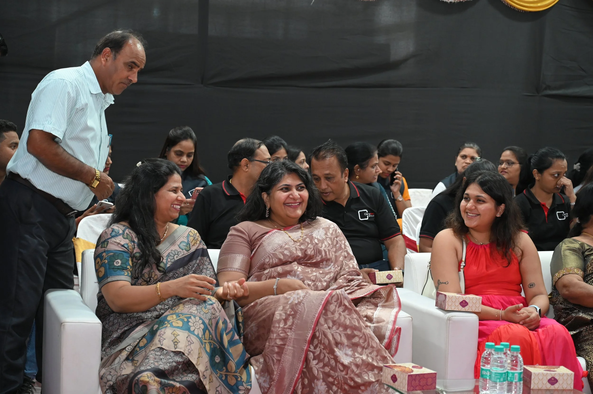 People sitting and engaging at a formal event, with some dressed in traditional Indian attire and others in casual shirts. They are smiling, talking, and looking at each other, with water bottles and gift boxes on the table in front.