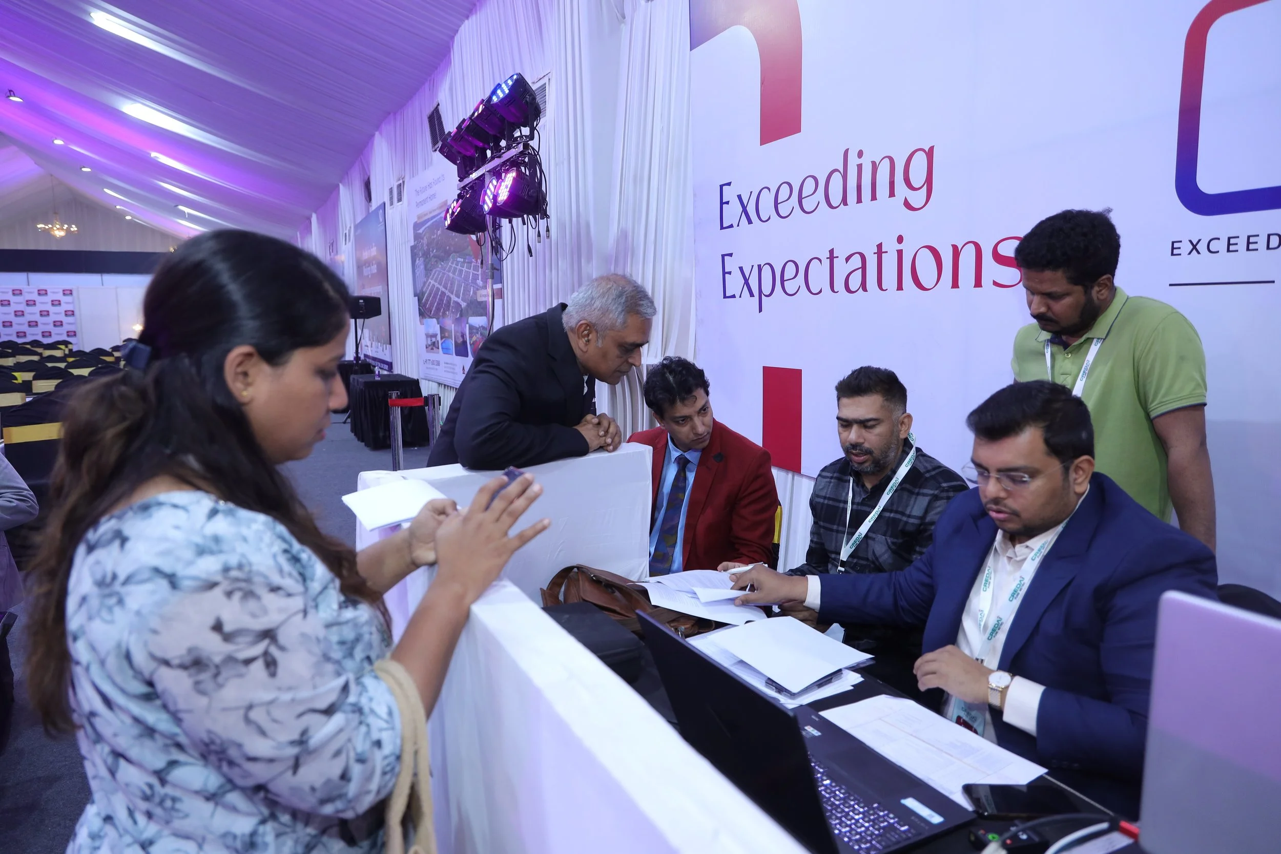 Group of people at a registration desk engaging in conversation and review of documents at a conference, with a backdrop displaying 'Exceeding Expectations'.