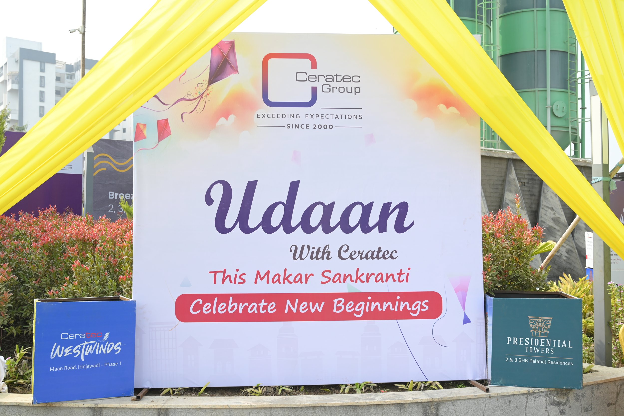 Large outdoor banner with the Ceratec Group logo, celebrating a new beginning for Makar Sankranti. The banner includes colorful graphics of kites flying in the sky, and the message: "Udaan With Ceratec - This Makar Sankranti Celebrate New Beginnings." There are two smaller signs at the bottom, one on the left promoting Ceratec Westwinds on Maan Road, Hinjewadi - Phase 1, and one on the right advertising Presidential Towers, 2 & 3 BHK Palatial Residences. The setup is decorated with yellow drapes overhead and plants at the base.