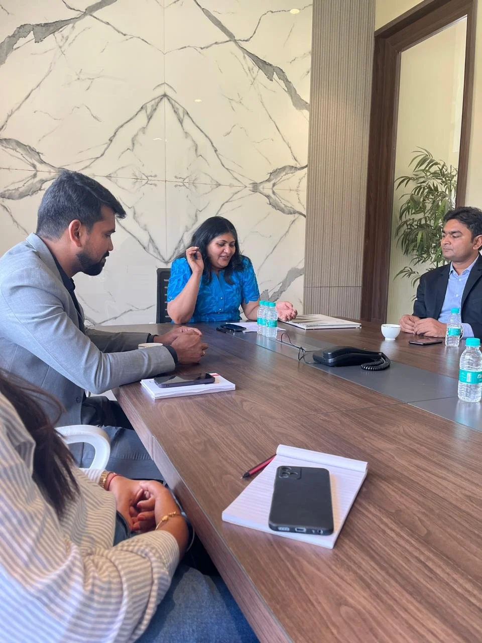 Four people seated around a conference table in a meeting room, with one woman speaking and three others listening, with notebooks, bottles of water, and a phone on the table.