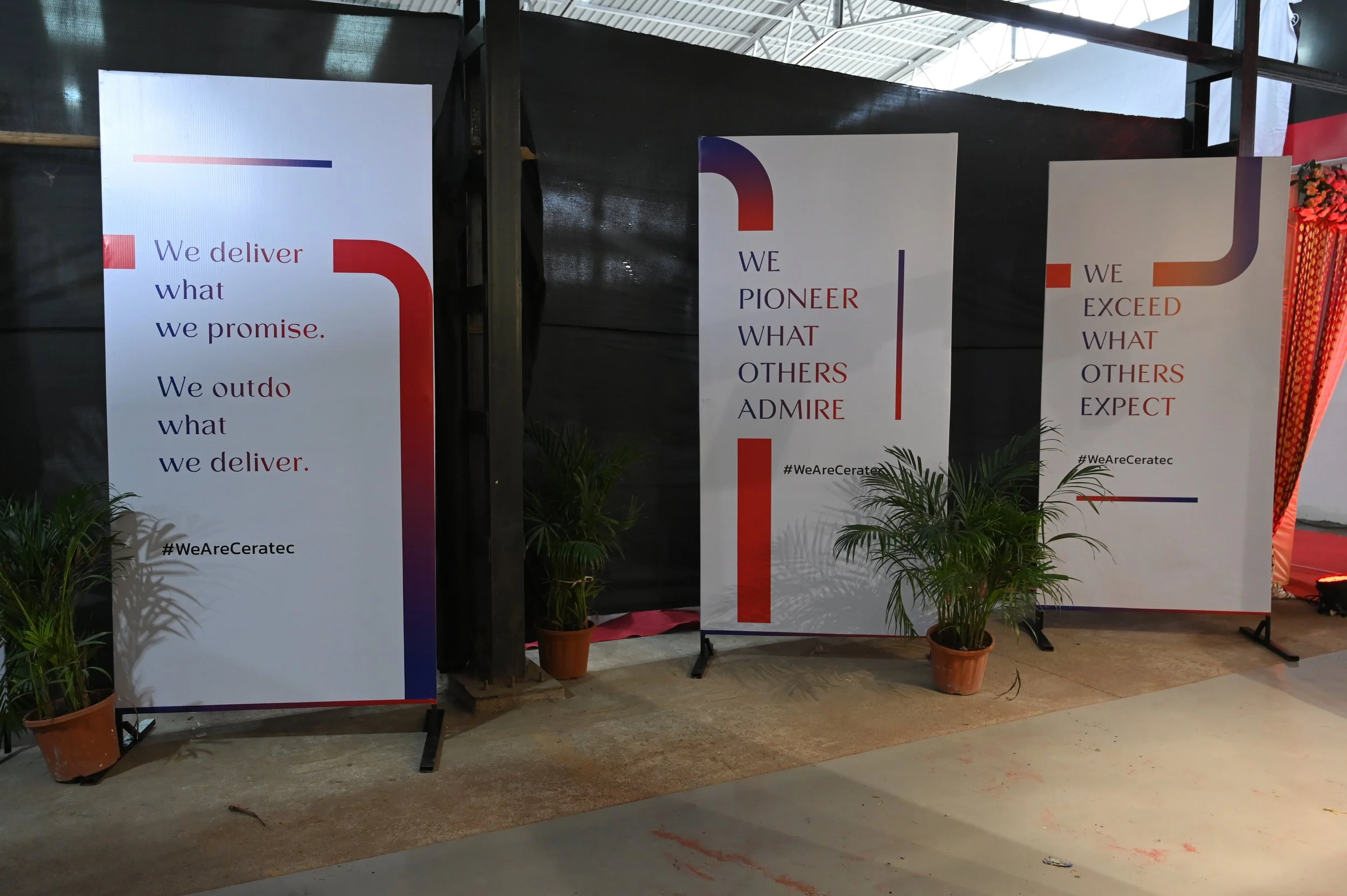 Three vertical banners with inspirational messages at a corporate or conference event. The banners have a white background with red, blue, and purple accents, and potted plants are placed in front of each. The left banner reads, 'We deliver what we promise. We outdo what we deliver.' The middle banner says, 'We PIONEER what others ADMIRE.' The right banner states, 'We EXCEED what others EXPECT.' All banners include the hashtag #WeAreCeratec.