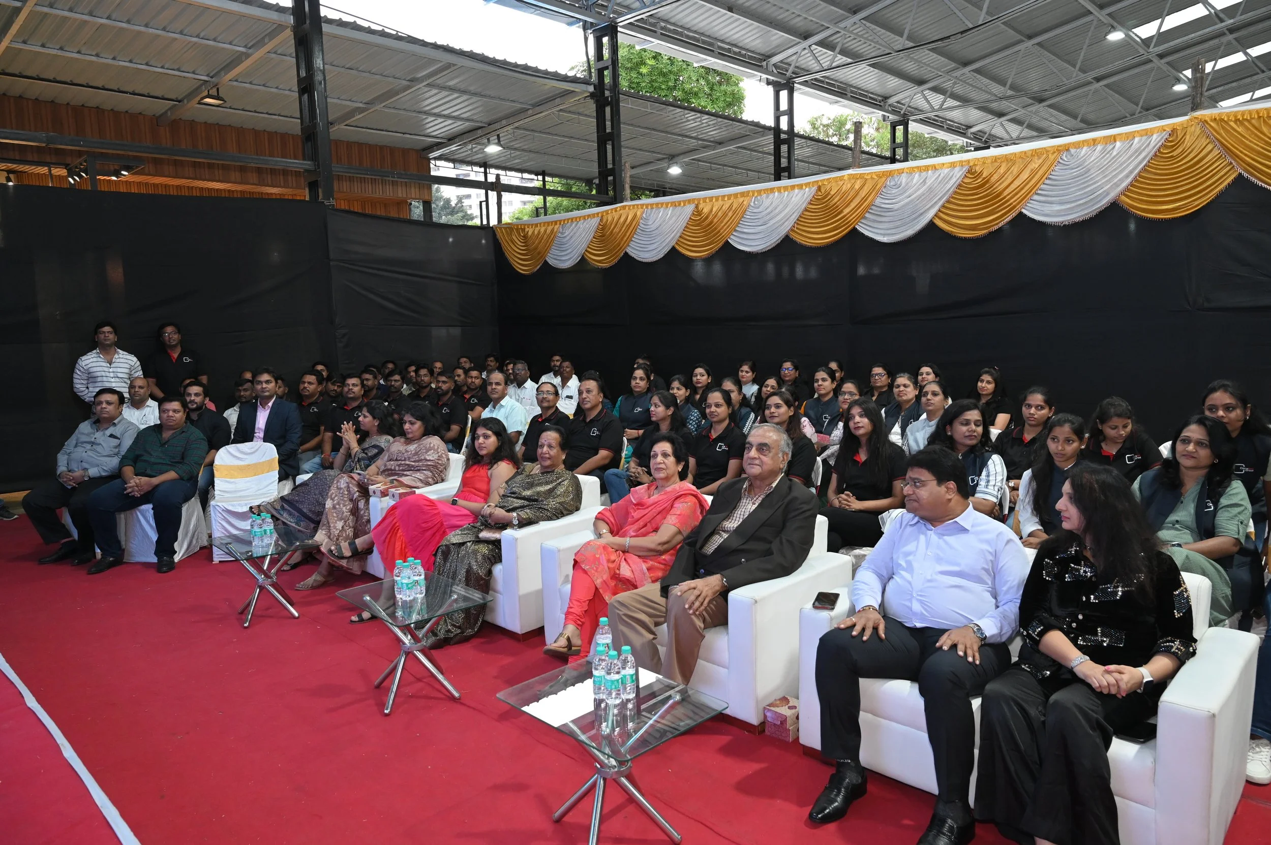 A large group of people seated in an indoor event space, with some seated on white chairs and others on black chairs, facing forward, under a metal roof with decorative yellow and white curtains.