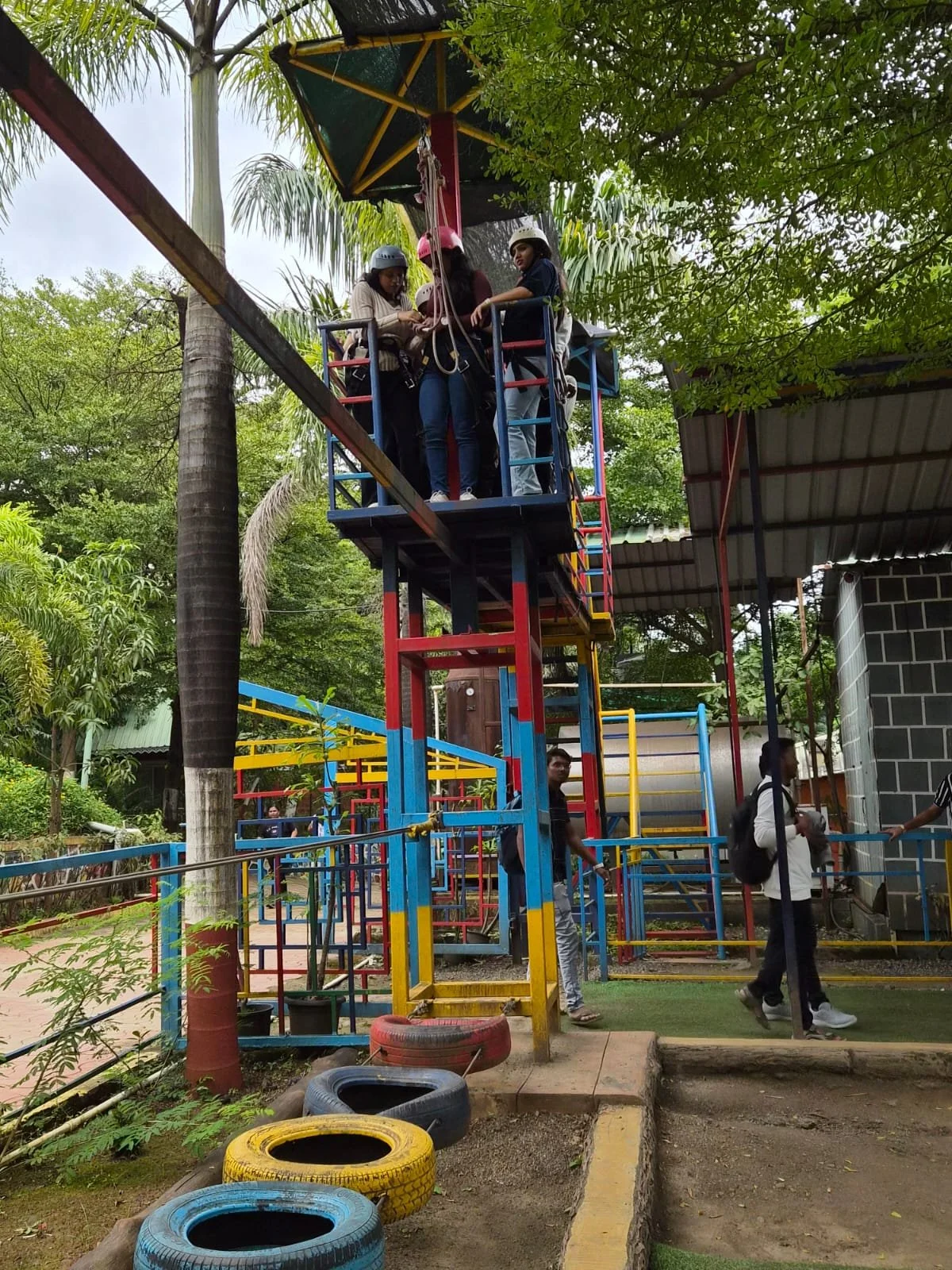 Children and adults standing on a colorful outdoor playground structure with a tall tower, surrounded by green trees and tires painted in bright colors on the ground.