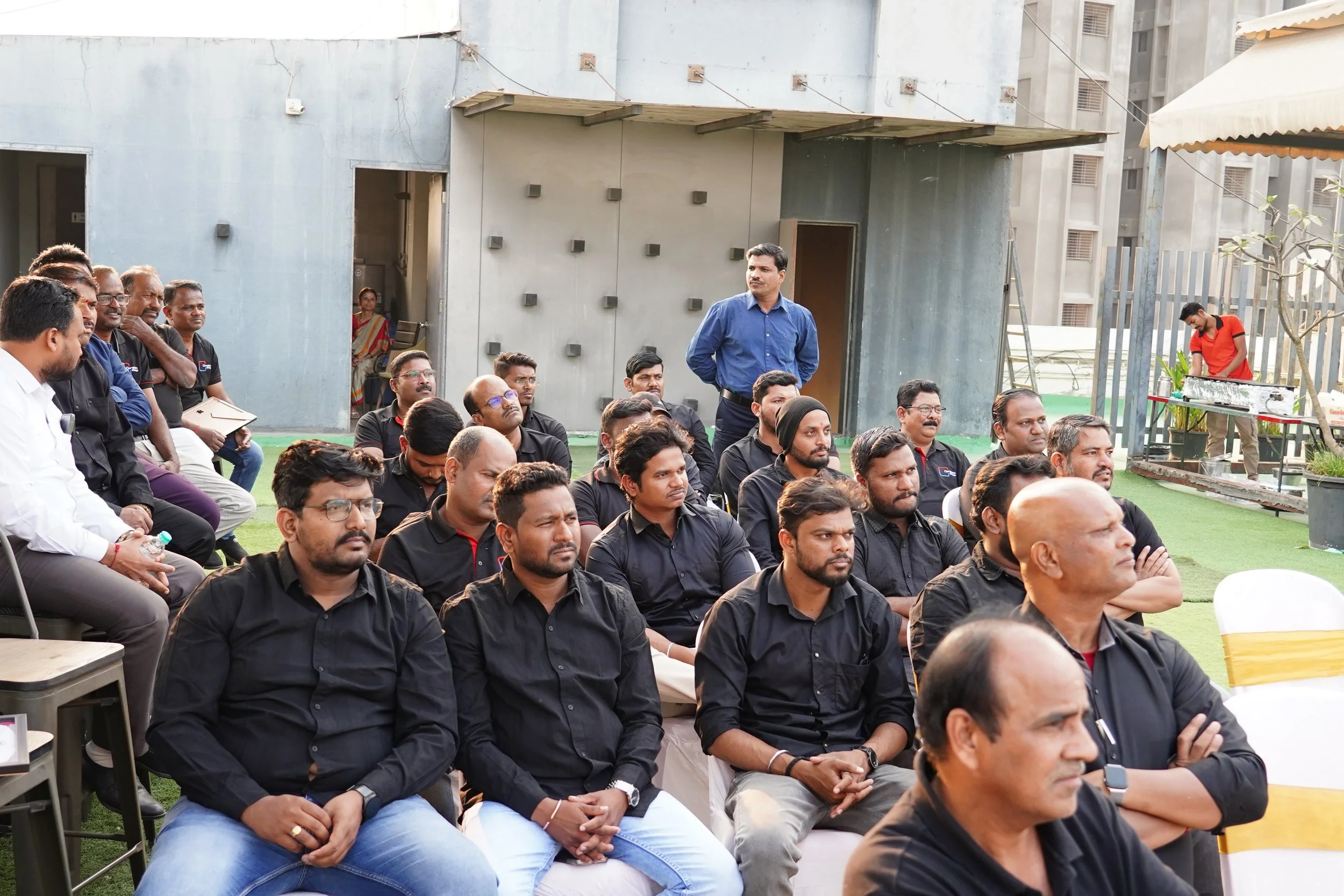 Group of men seated outdoors at an event, many wearing black shirts, with some standing in the background, and a woman in traditional attire visible through an open doorway.