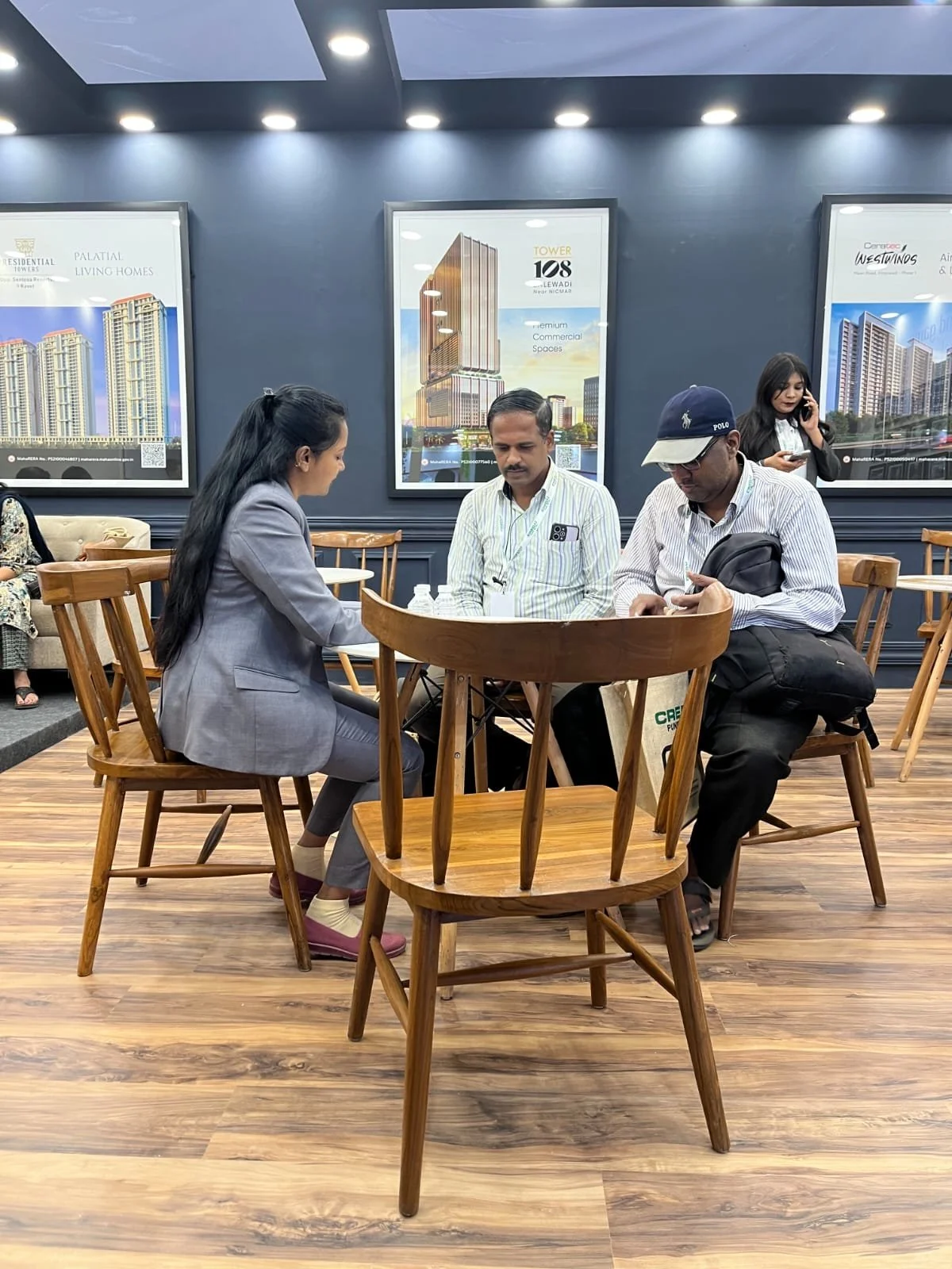 Three people sitting at a round wooden table in a modern indoor setting with large posters of building plans on the wall behind them. A woman with long black hair in a gray suit is speaking with two men, one in a white striped shirt and the other in a striped shirt with a cap, both looking at their phones. A woman in the background is talking on her phone.