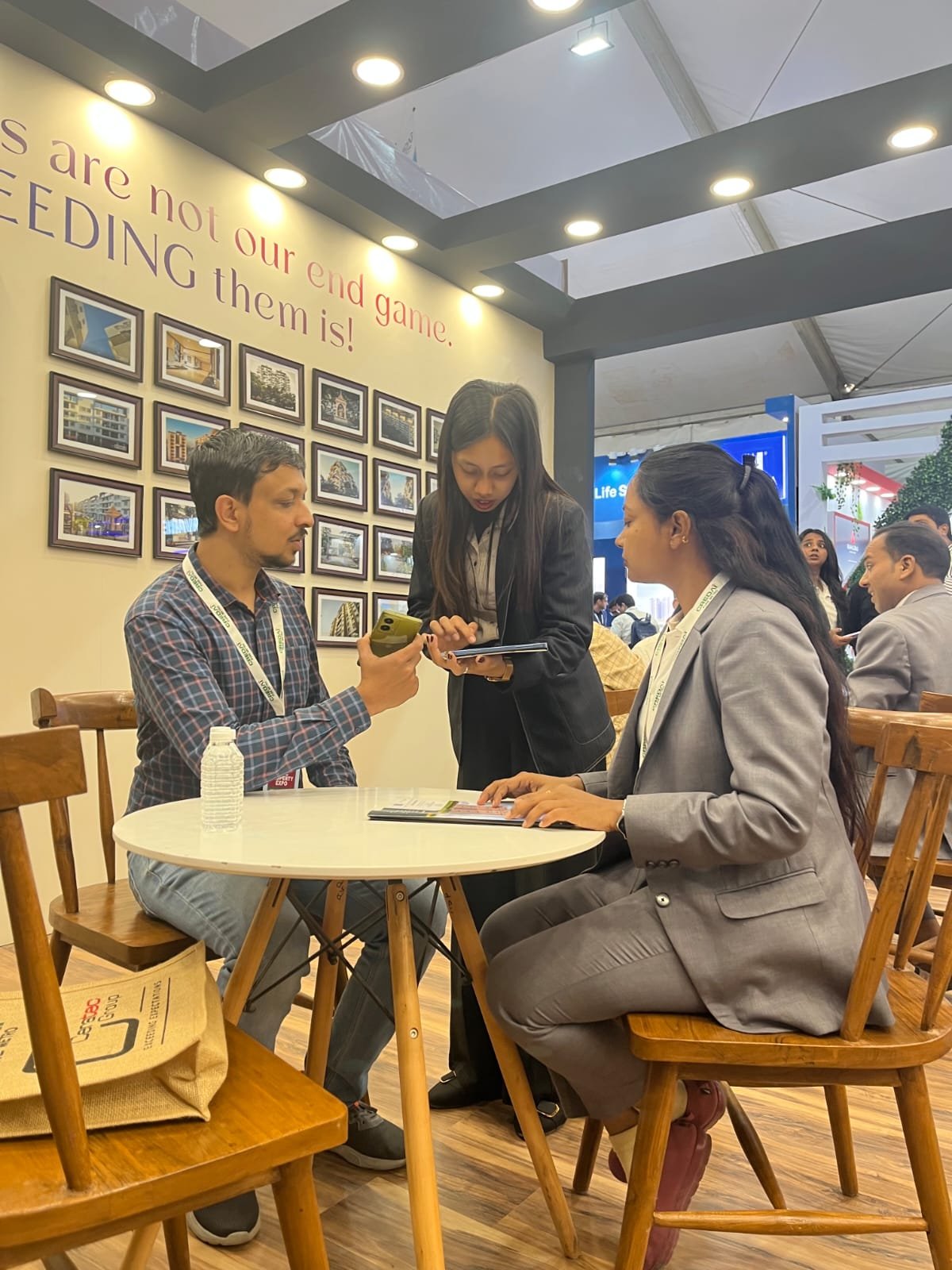 Three people engaged in a conversation at a round white table in a busy indoor setting. One woman is standing, showing something on a tablet to a man and a woman seated. The seated woman is wearing a gray suit, and the man is dressed casually. The background has framed pictures on a wall and a slogan that reads 'We are not our end game, we are editing them is!'.