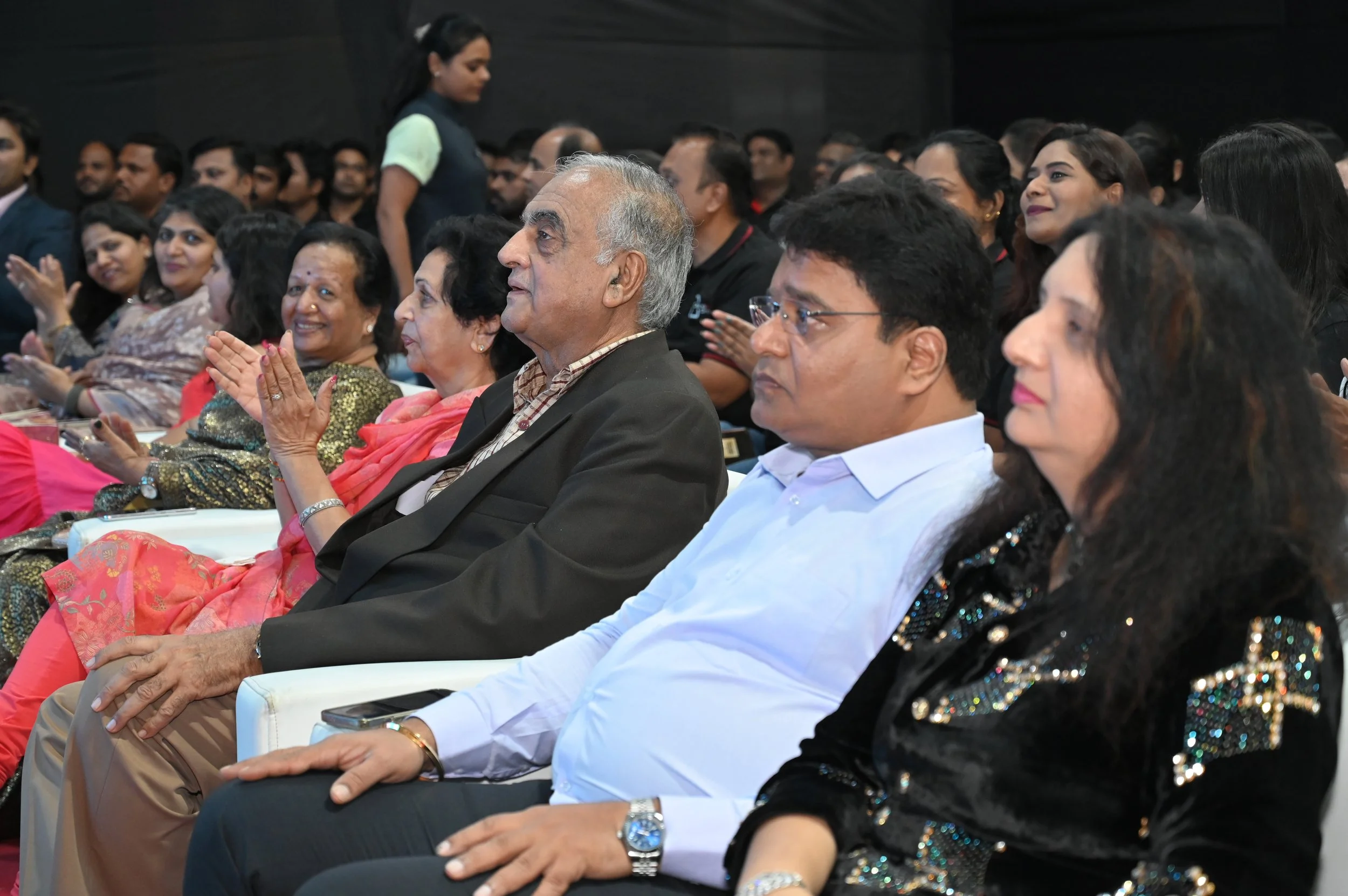Audience members sitting in chairs during a formal event, some clapping and attentively listening.