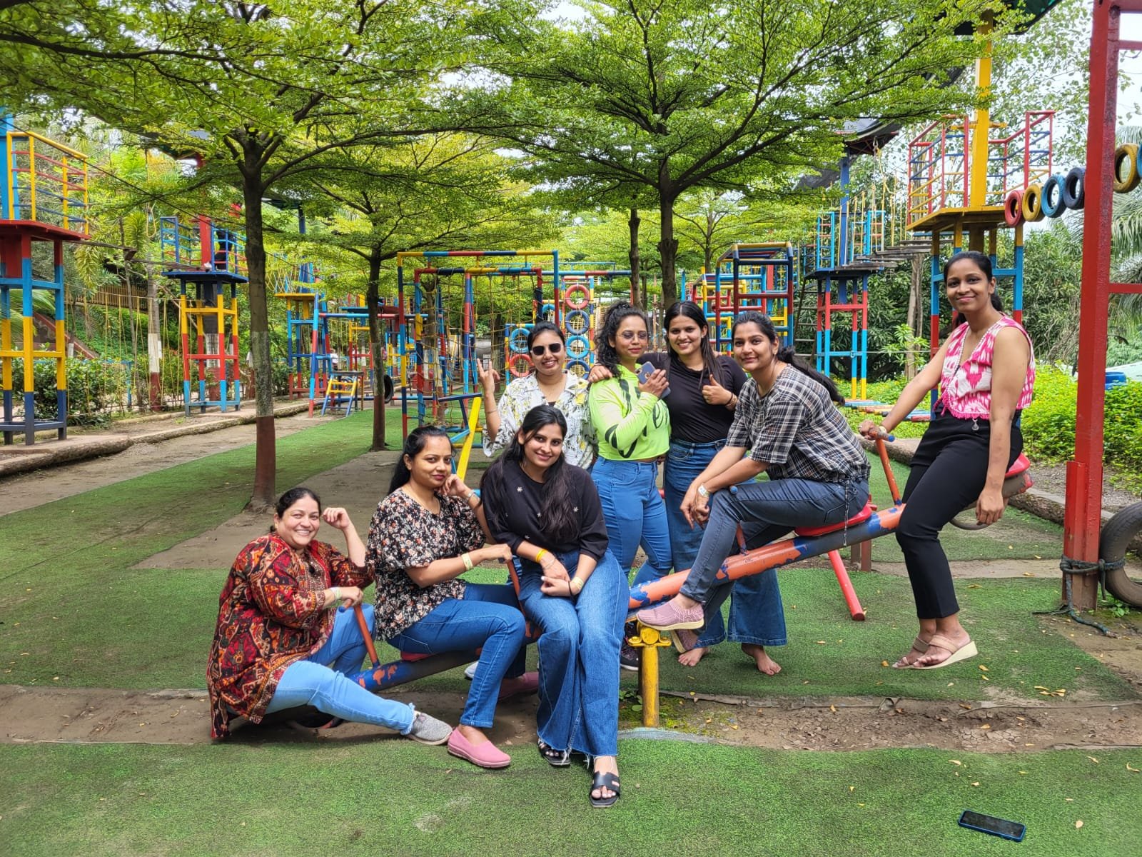 A group of nine women smiling and posing at an outdoor playground, surrounded by colorful play structures and green trees.