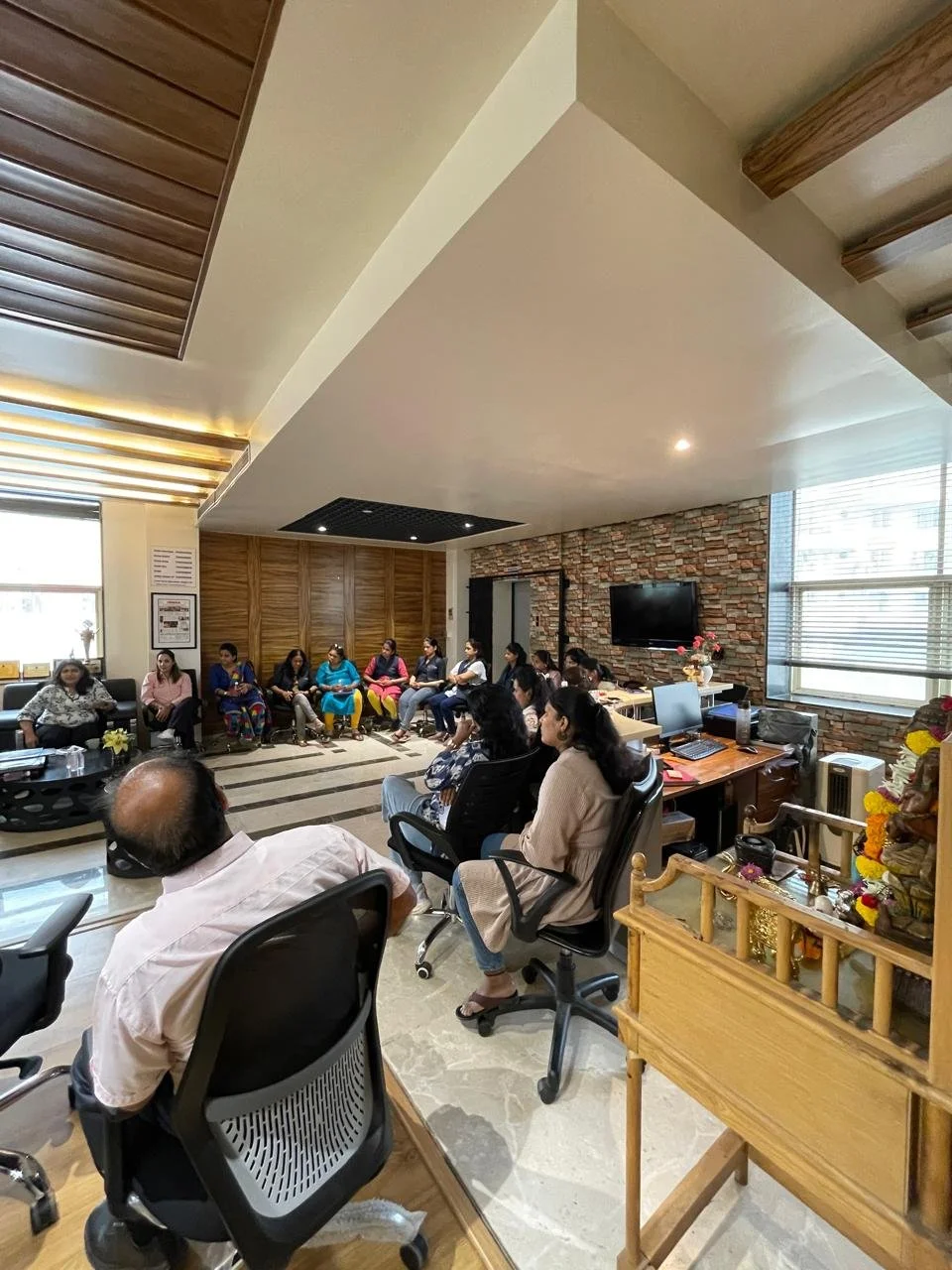 Group of people sitting in a conference room, some on chairs and some on a sofa, listening to a speaker. The room has windows, a brick wall with a mounted TV, and wooden accents.