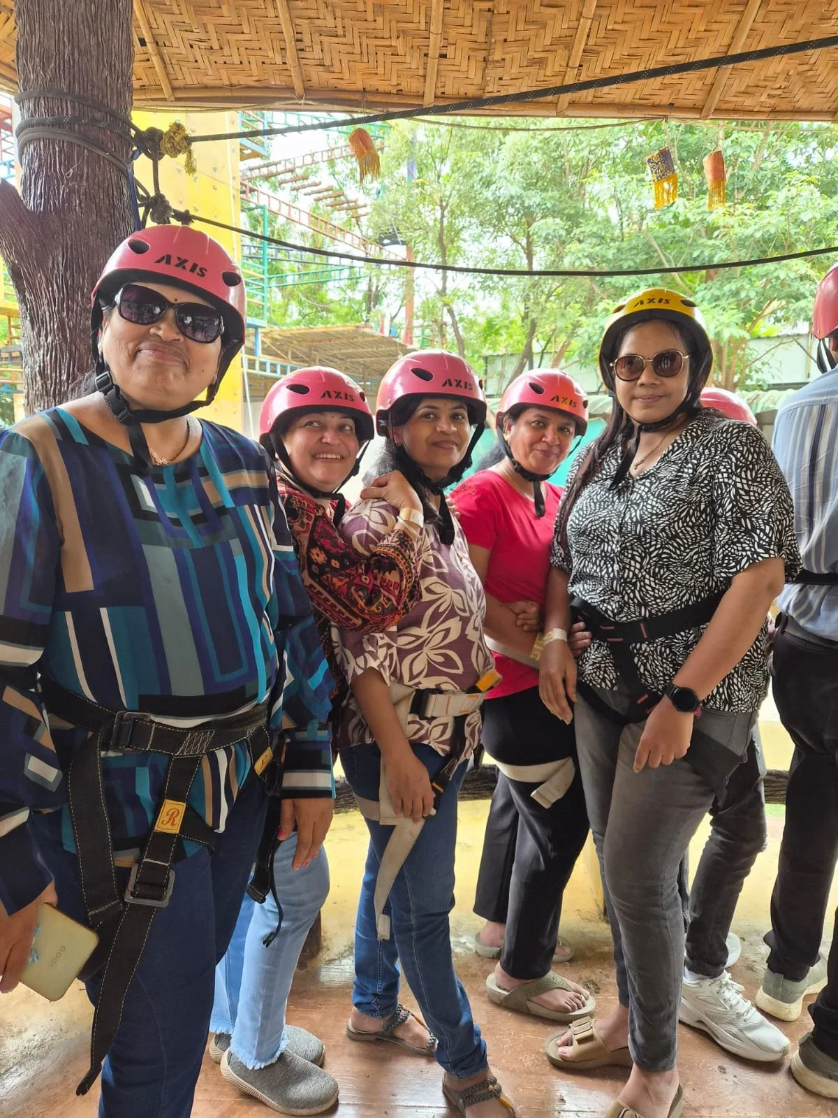 A group of women wearing safety helmets and casual clothing, standing together outdoors under a thatched roof, ready for an outdoor activity.