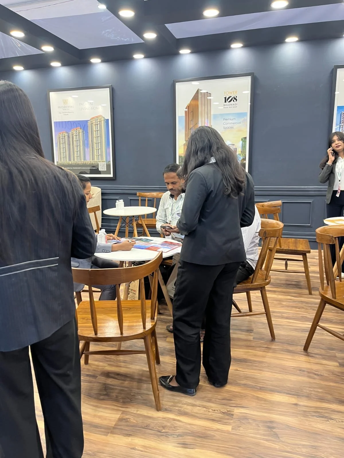 Business meeting with people in formal attire gathered around a table in a modern, well-lit room with blue walls, wood flooring, and framed architectural posters.