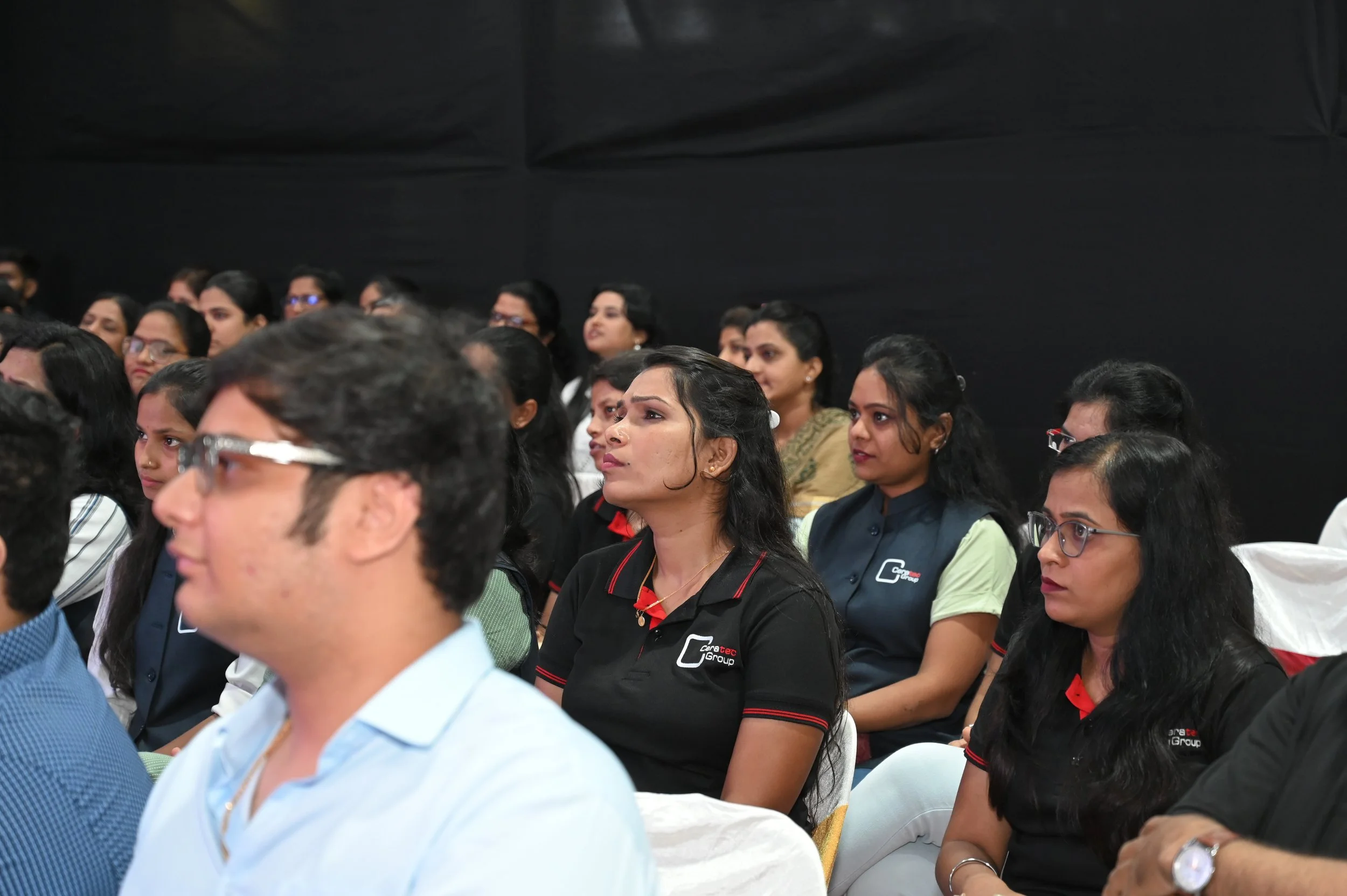 Allergic audience listening attentively at a conference or seminar, seated in rows against a black backdrop, with some wearing branded shirts.