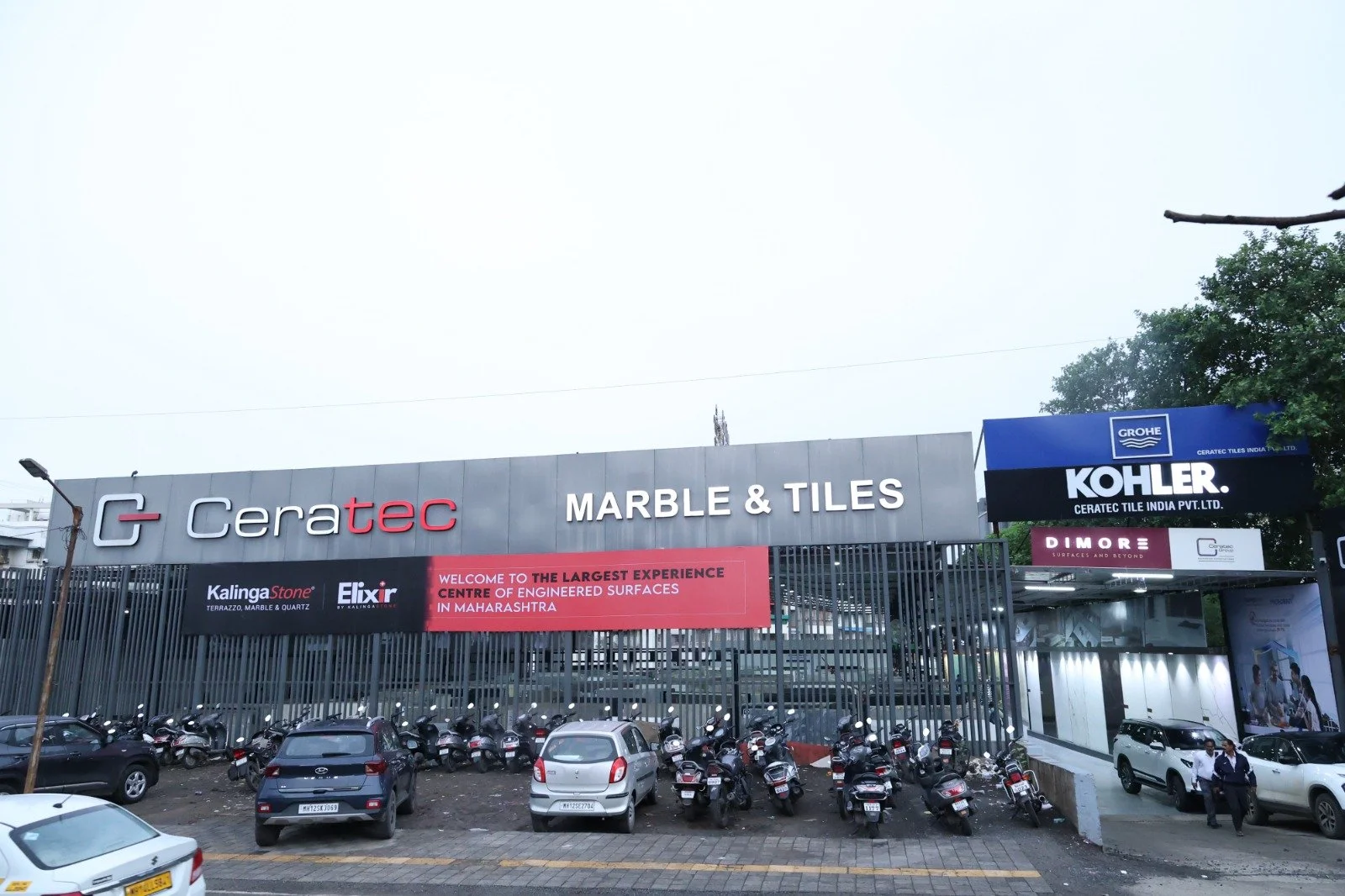Exterior view of a marble and tiles store with parked cars and bikes in front, showing signs for Ceratec, KalingaStone, Elixir, Grohe, Kohler, and Dimore. Ceratec Marble and Tiles Showroom.