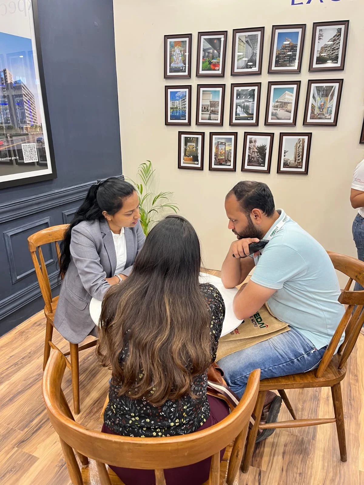 Three people sitting around a table in a restaurant or cafe, with a wall featuring framed pictures of buildings and cityscapes behind them.