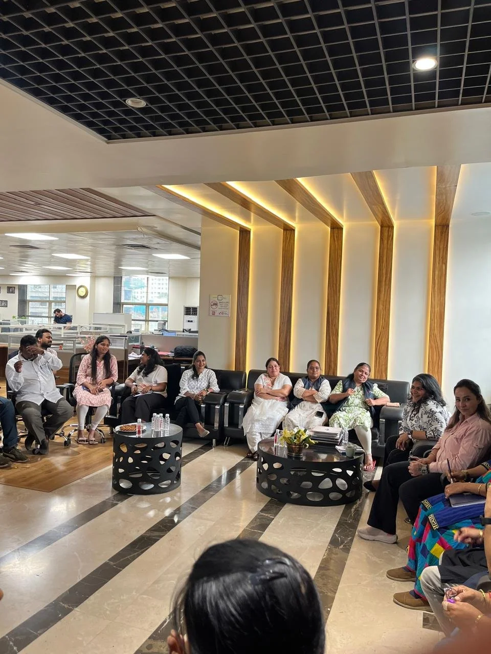 Group of people sitting on black sofas in a modern office lounge, engaging in discussion or waiting, with some bottles of water and magazines on small tables.