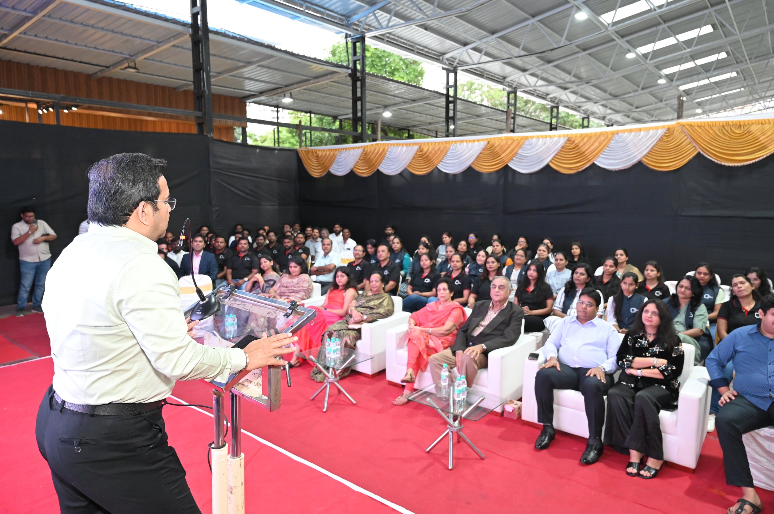 A man in light-colored shirt speaking at a podium to a large audience in a spacious indoor venue with a high ceiling, decorated with gold and white drapes.