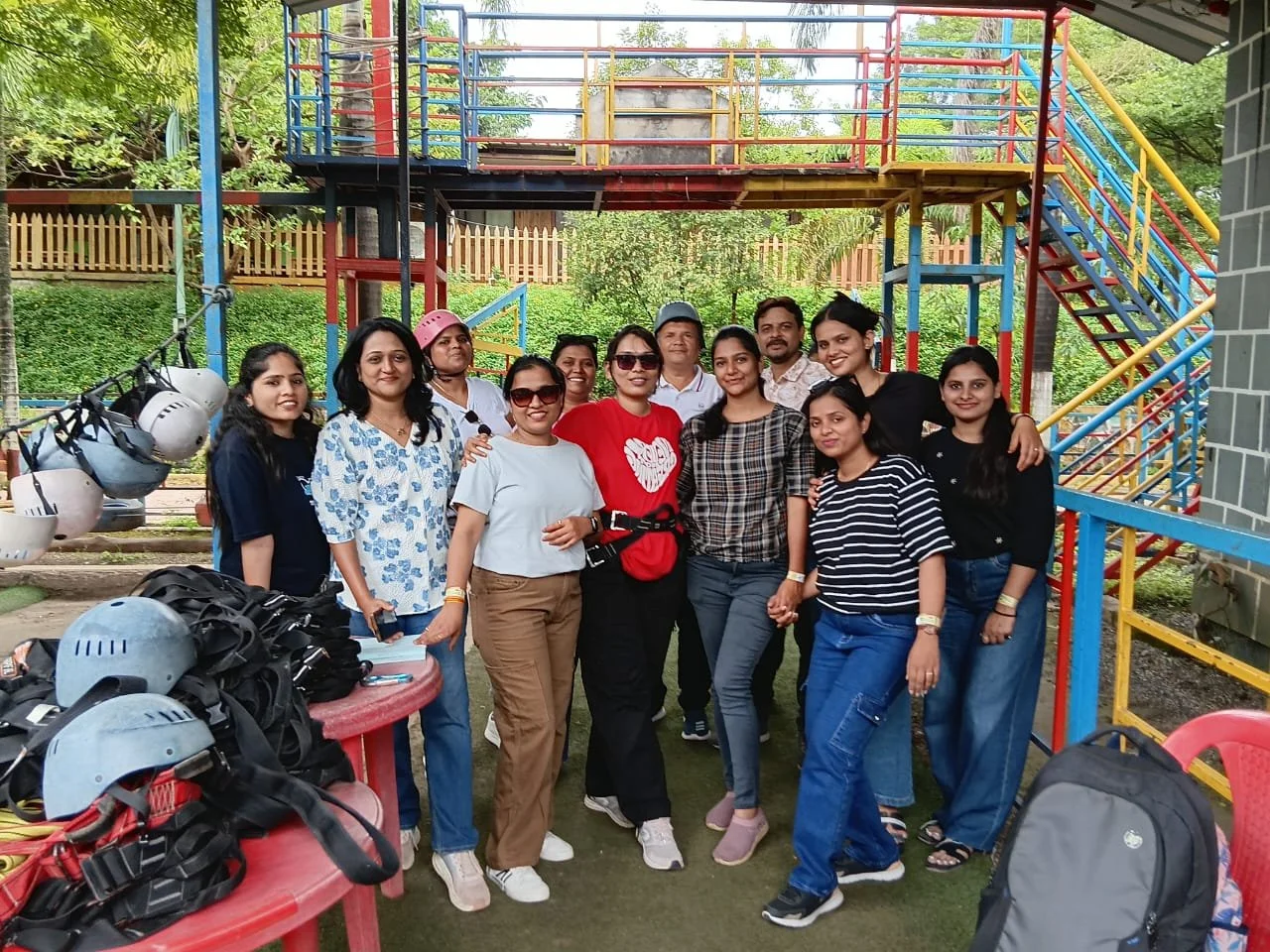 A group of twelve people posing together outdoors in front of colorful playground structures. The group includes men and women smiling at the camera, some wearing sunglasses and casual clothing. Helmets and backpacks are placed on a red table in the foreground.