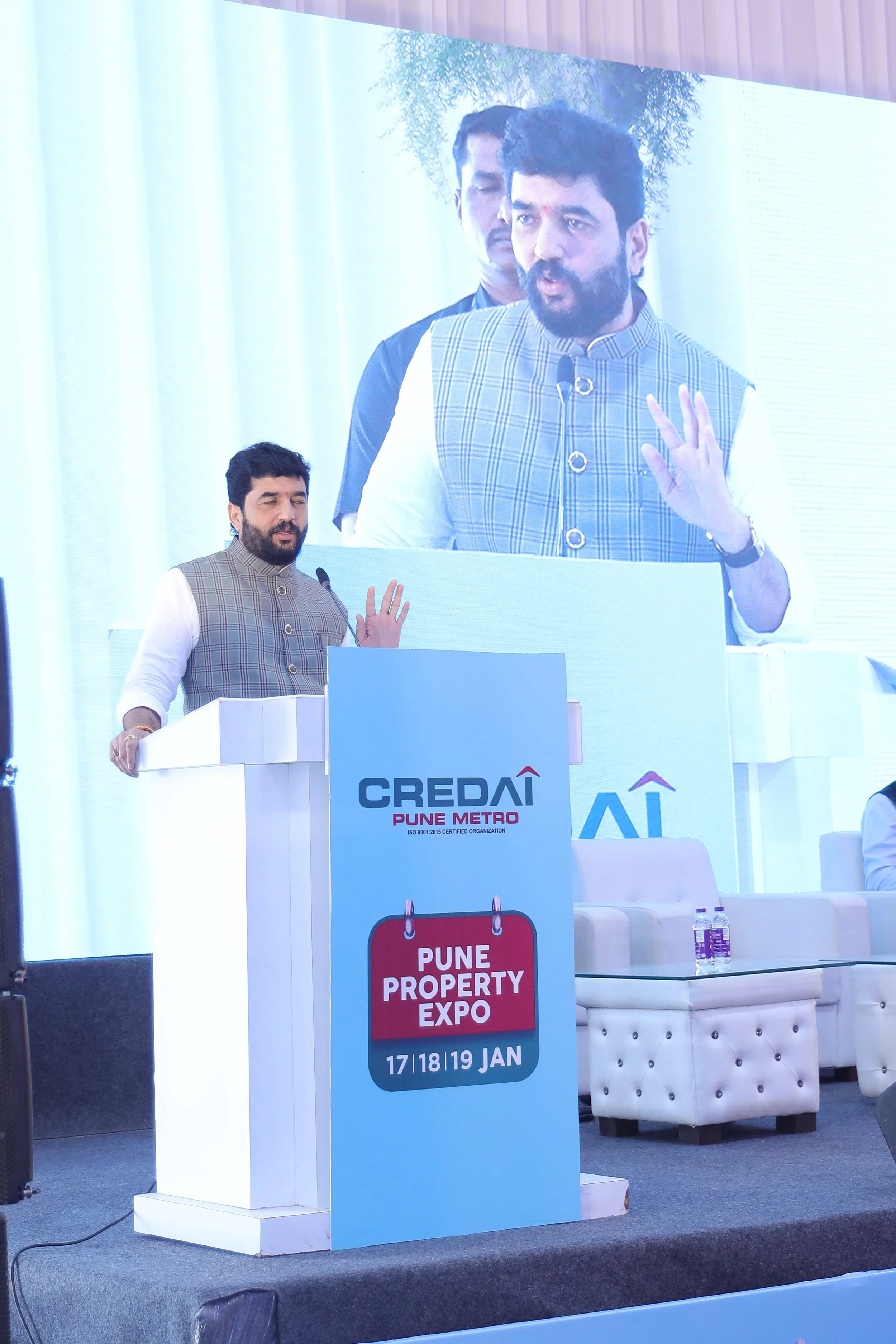Man speaking at podium during Pune Property Expo, with large screen behind him displaying his image.