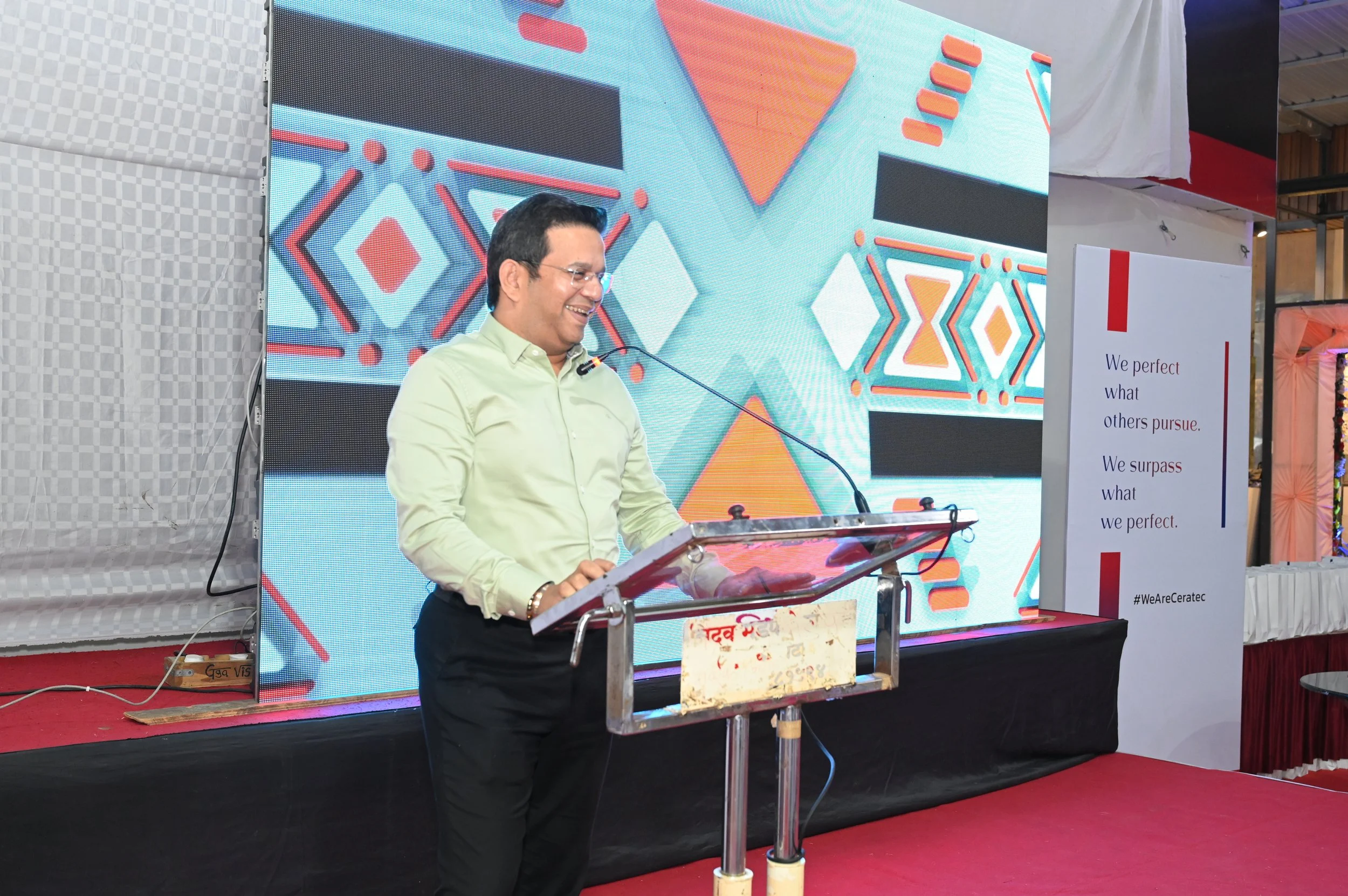 A man in a light green shirt and black pants speaking at a podium, smiling, with a large colorful geometric patterned screen behind him and a white sign with red and blue text on his right.