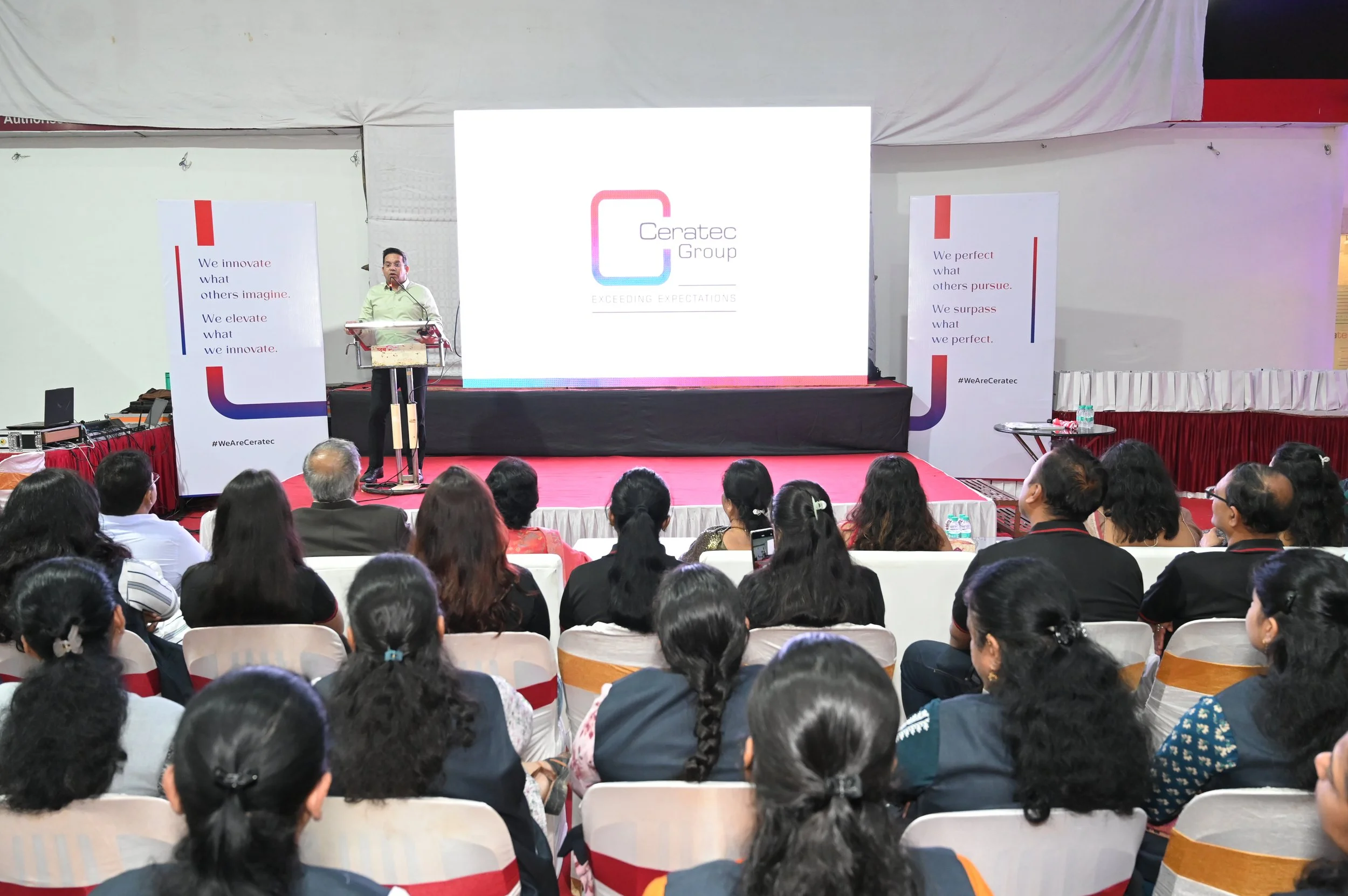 A speaker on stage at a conference presenting to an audience seated in rows. The stage features a large screen with the Ceratec Group logo displayed. Two vertical banners with company slogans and hashtags flank the stage. The audience appears engaged, with some taking notes or photos.