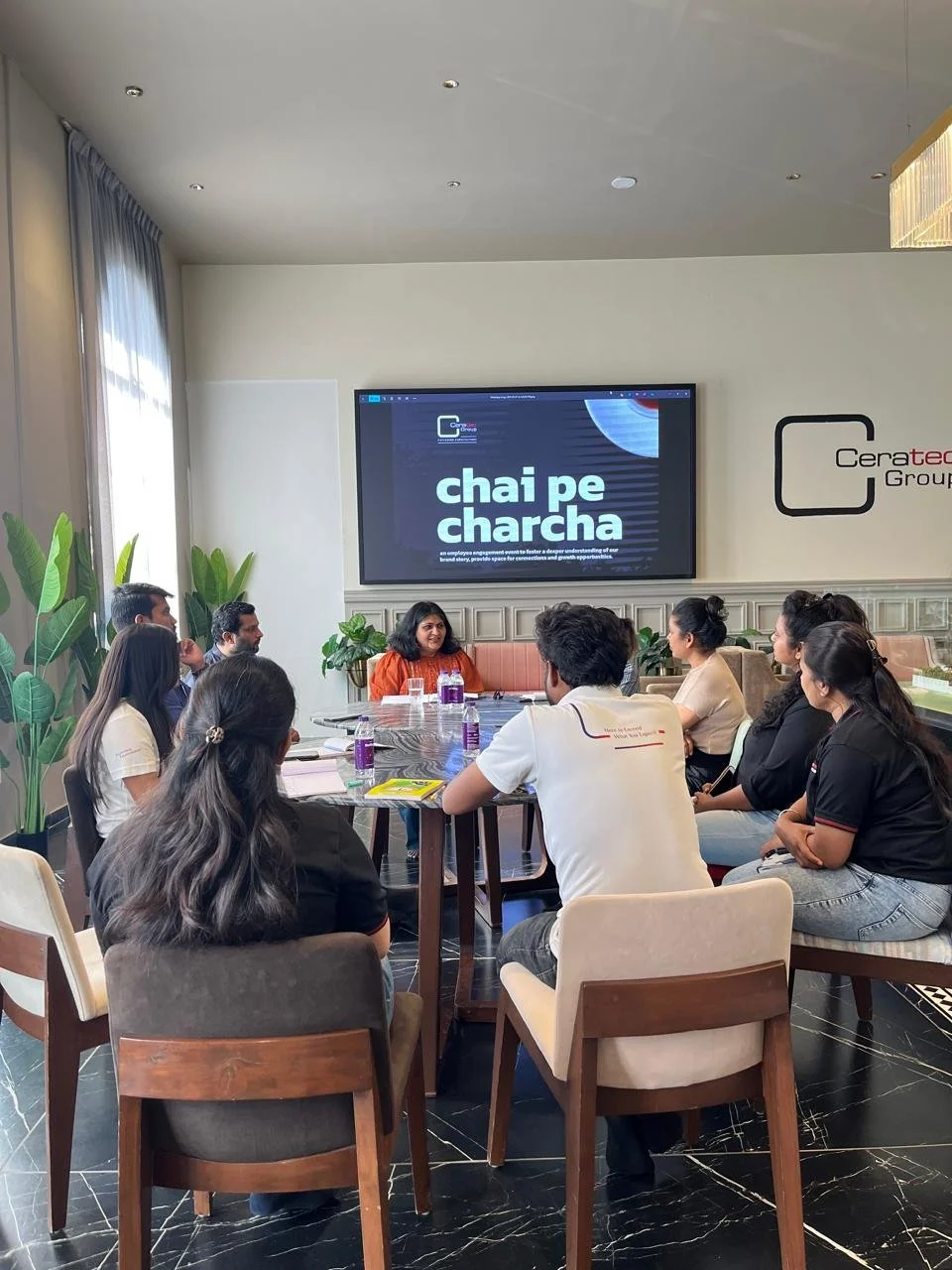 Group of people attending a meeting or training session in a conference room, sitting around a large table, with a woman leading the session displayed on a large screen at the front of the room.