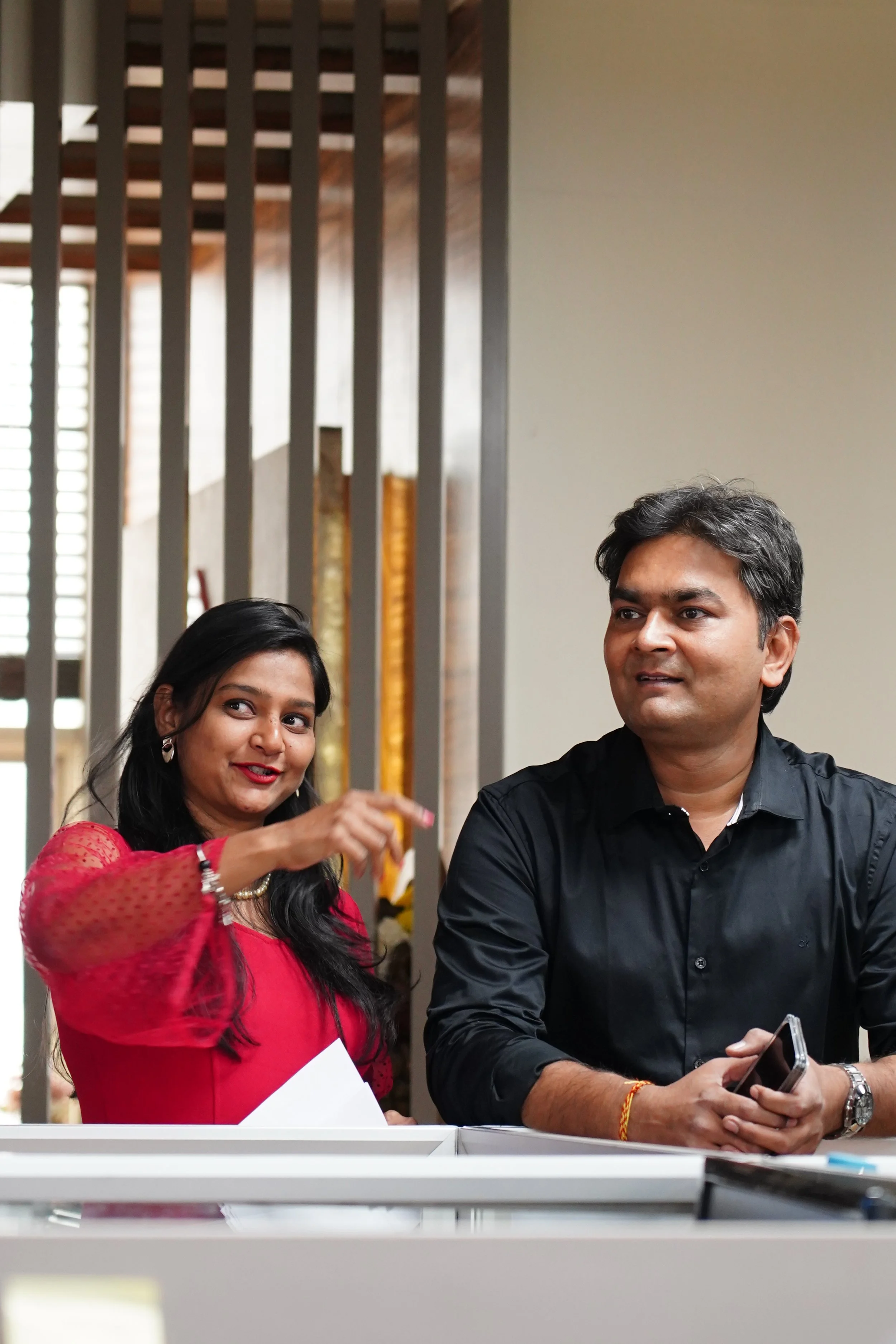 A woman in a red top and a man in a black shirt stand together indoors, engaging in conversation. The woman is gesturing with her hand and holding papers, while the man is holding a smartphone.