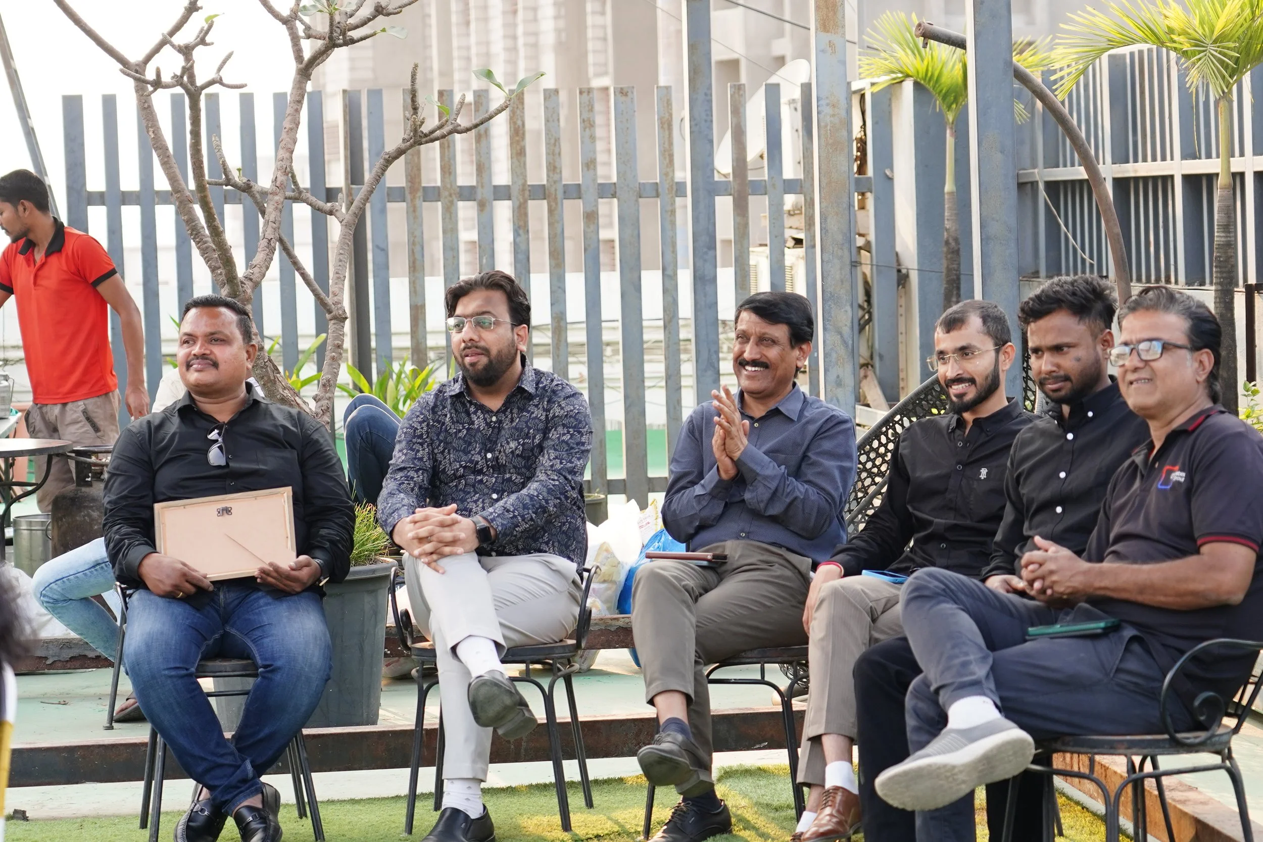 Group of six men sitting outdoors near a metal fence, smiling and clapping, with a tree and potted plants in the background.