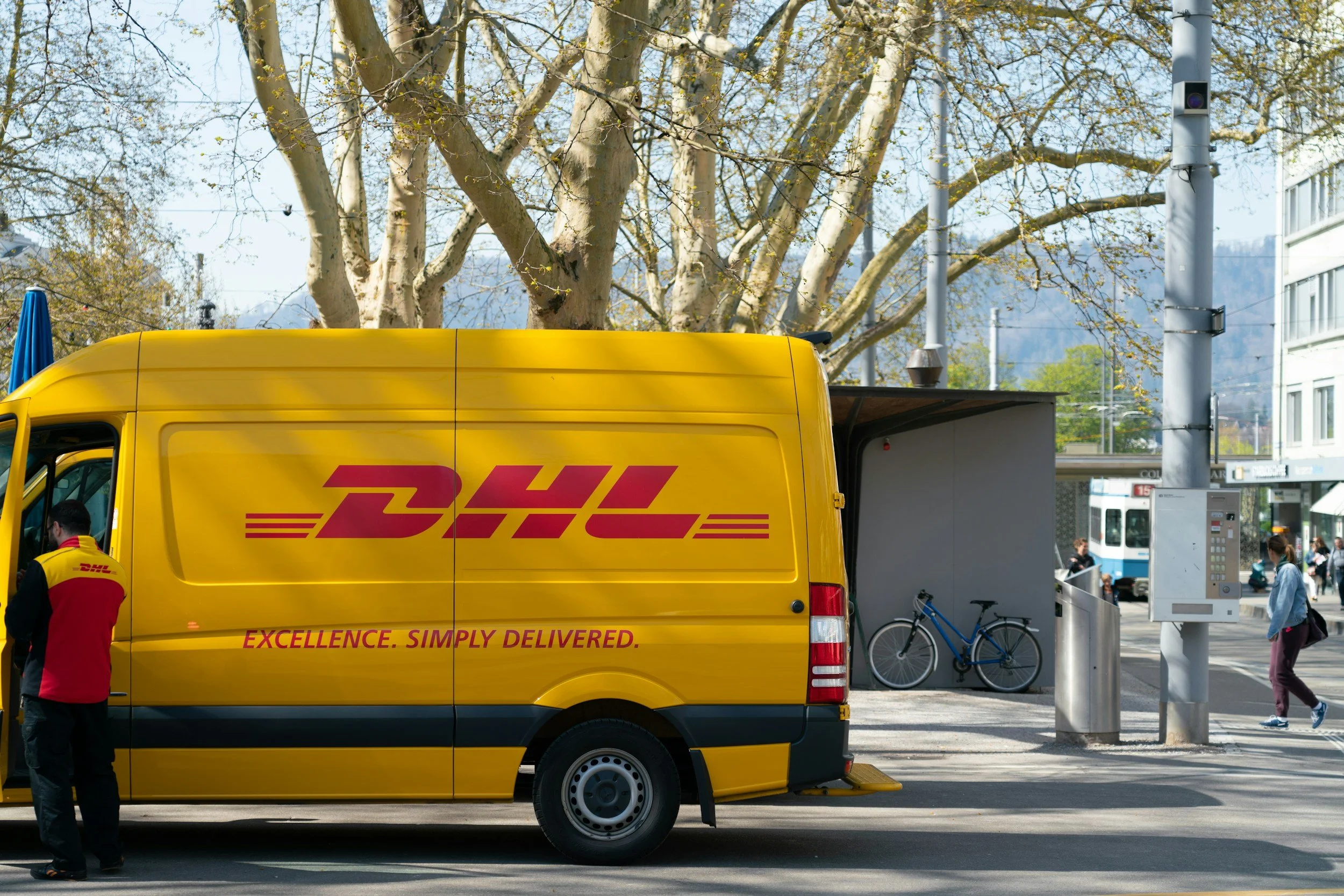 Yellow DHL delivery van parked on city street with delivery person standing beside it, a blue bicycle parked nearby, pedestrians walking, trees, and a tram in the background.