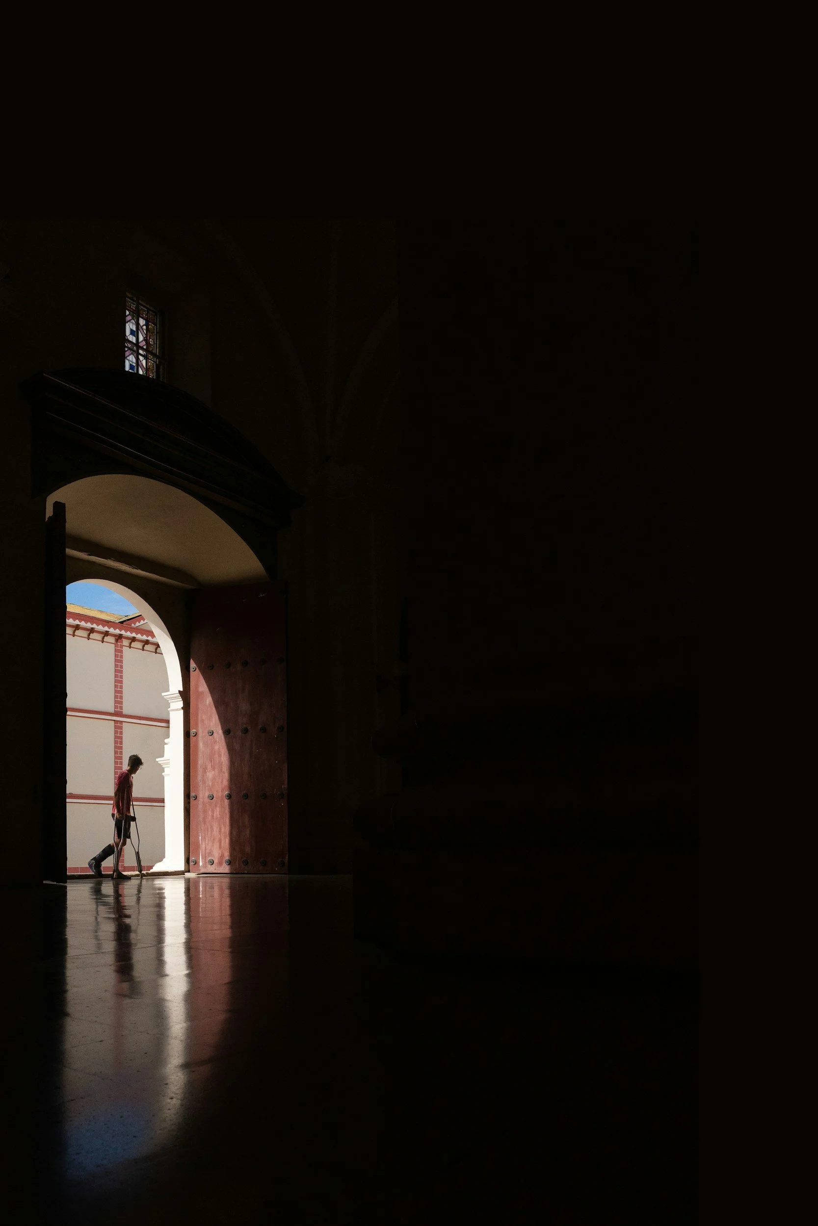 A person with a dog walking through a large arched doorway inside a dimly lit building, with sunlight illuminating the doorway and the person.