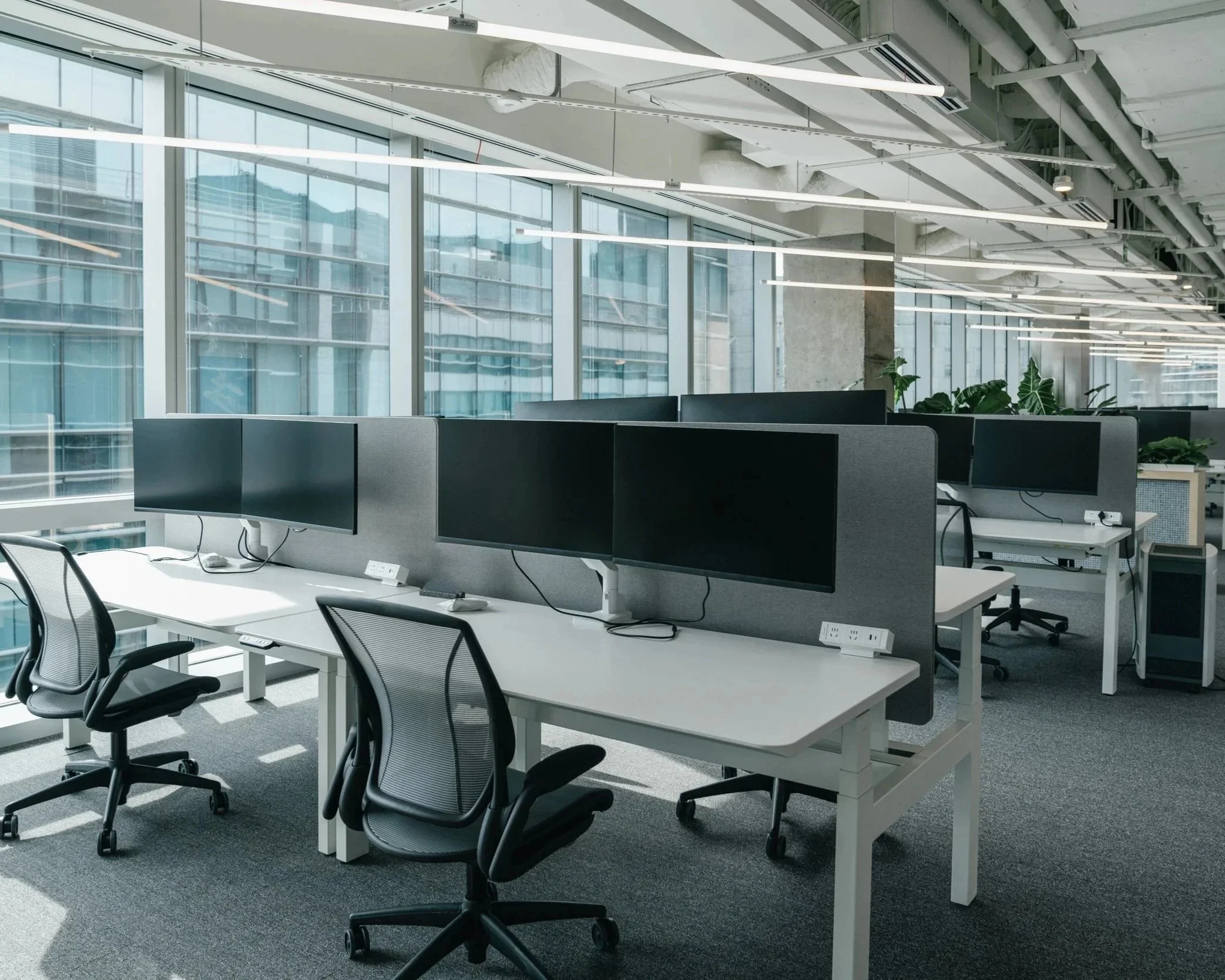 Modern office workspace with multiple desks, drop down lighting, ergonomic chairs, and dual monitors, alongside large windows letting in natural light.