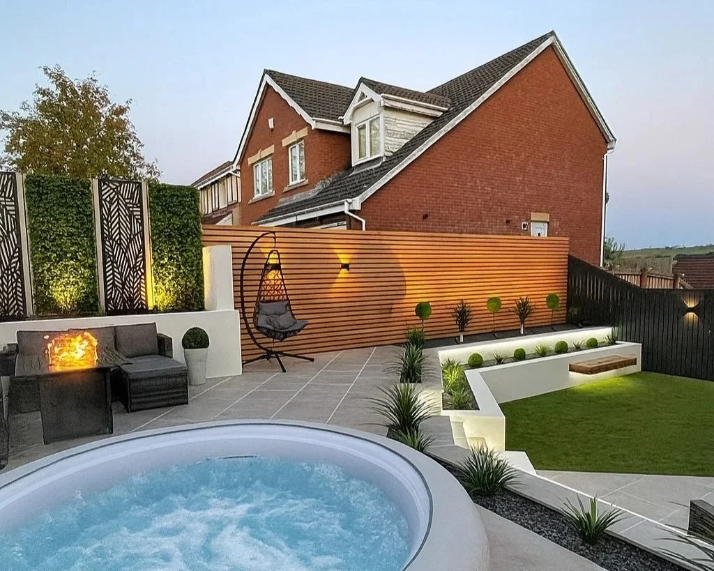 Residential backyard with hot tub, outdoor seating, outdoor lighting, decorative plants, wood privacy fence, and a lawn area, with a brick house in the background.
