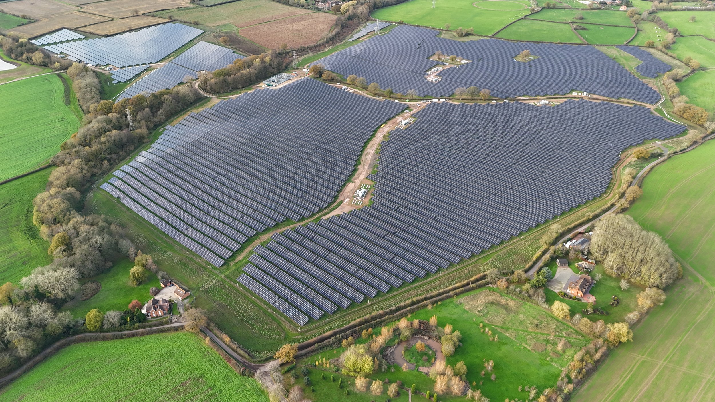 An aerial view of a solar farm with numerous solar panels installed on rolling green fields, surrounded by trees, roads, and a few houses.