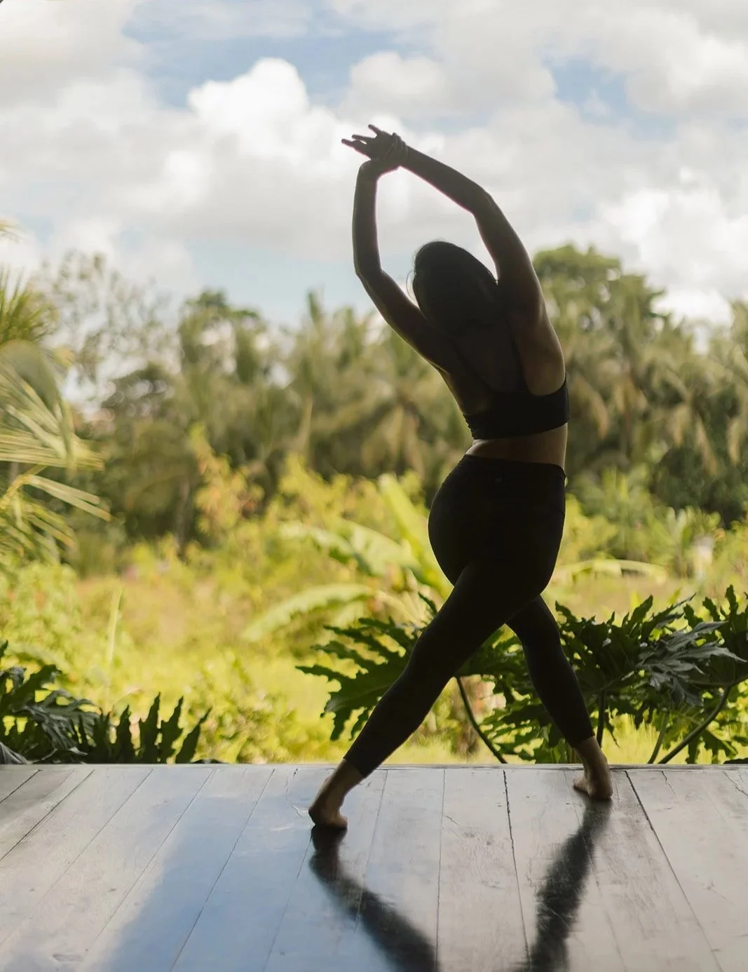 A woman practicing yoga outdoors on a wooden deck, striking a pose with arms overhead and one leg bent, silhouetted against a lush green landscape and cloudy sky.