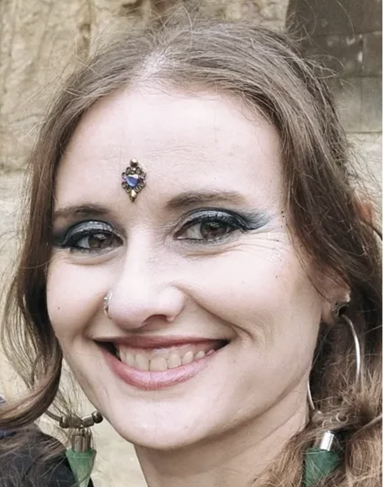 Retrato de una mujer con maquillaje llamativo, joyería en la frente y pendientes grandes, sonriendo.